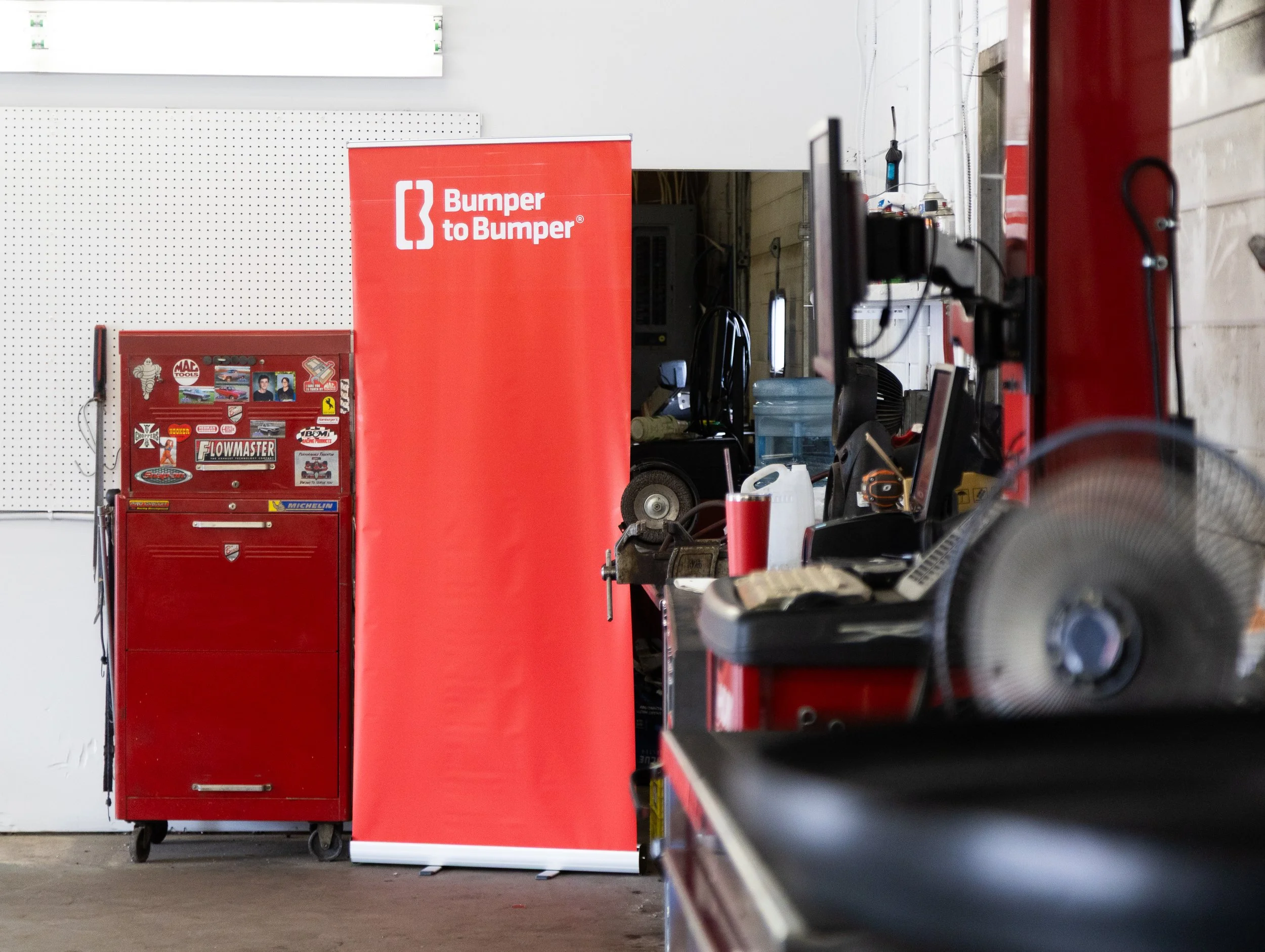Inside a garage workshop with a red tool cabinet, a red banner with 'Bumper to Bumper' logo, and various tools and equipment on workbenches and shelves.