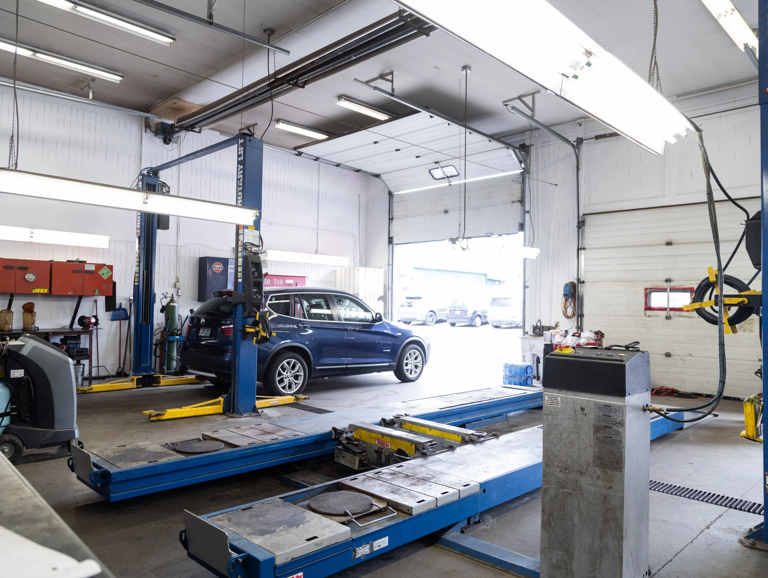 Interior of an automotive repair shop with a car lift, a black SUV on the lift, and equipment around the garage.