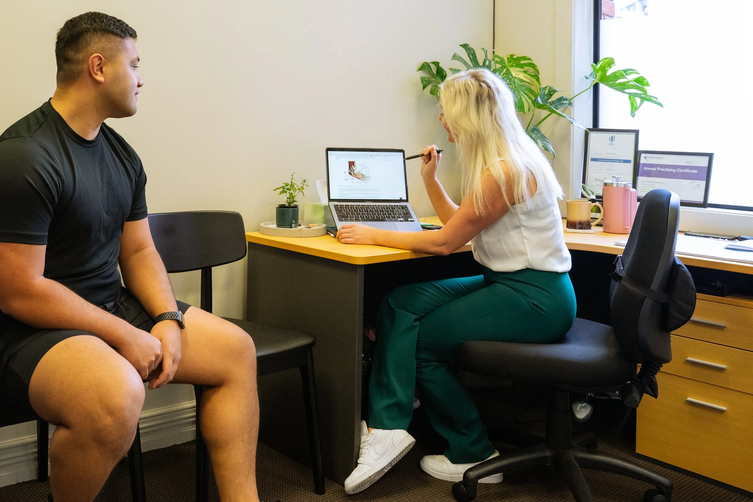 A healthcare professional sitting at a desk consulting with a male patient who is seated across from her in an office with potted plants and framed certificates.