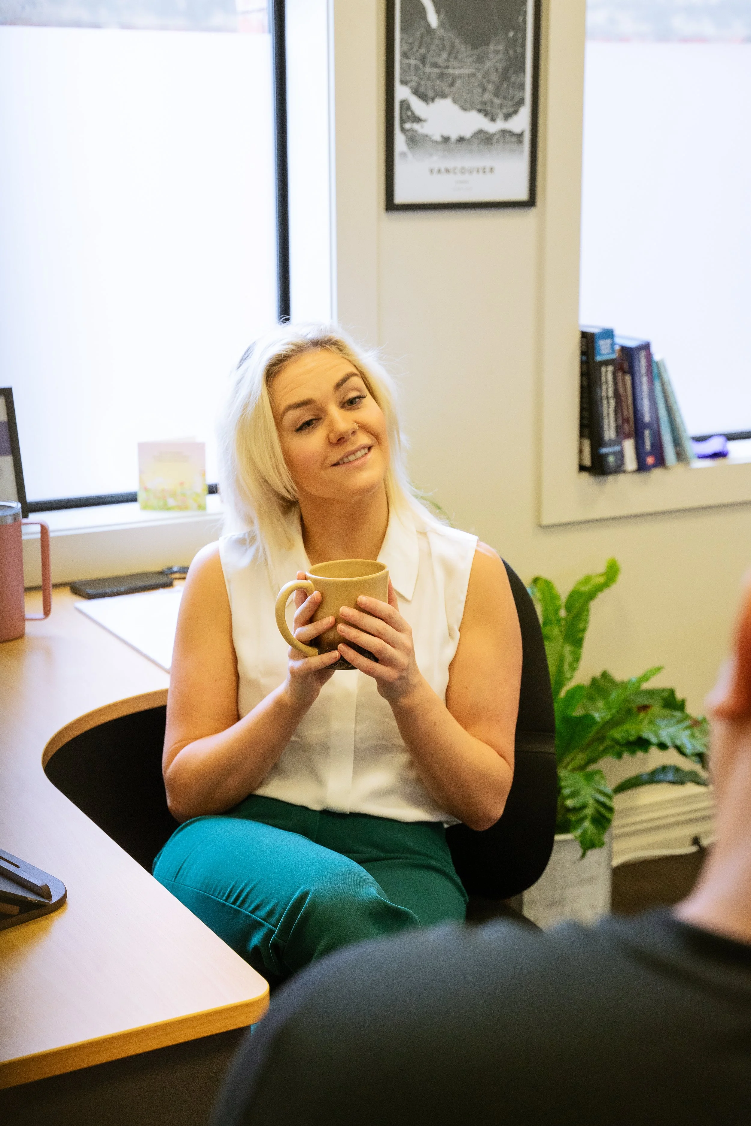 A woman with blonde hair sitting at a desk, smiling and holding a beige mug, engaged in conversation with a person whose back is visible.
