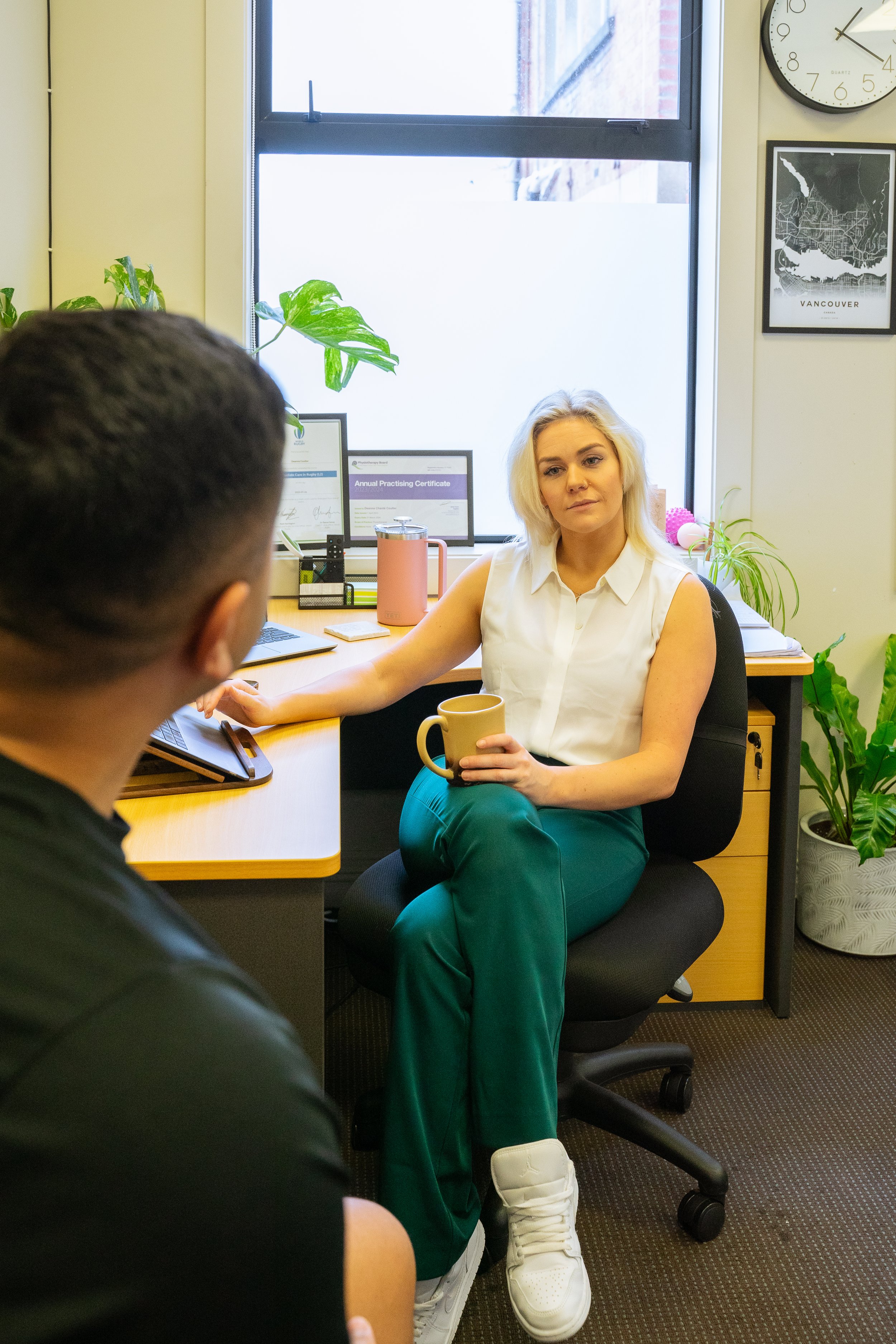 A woman with blonde hair sitting in an office chair, holding a mug, and listening to a man whose face is not visible, in a desk with plants and documents. The office has a window, a clock, and framed pictures on the wall.