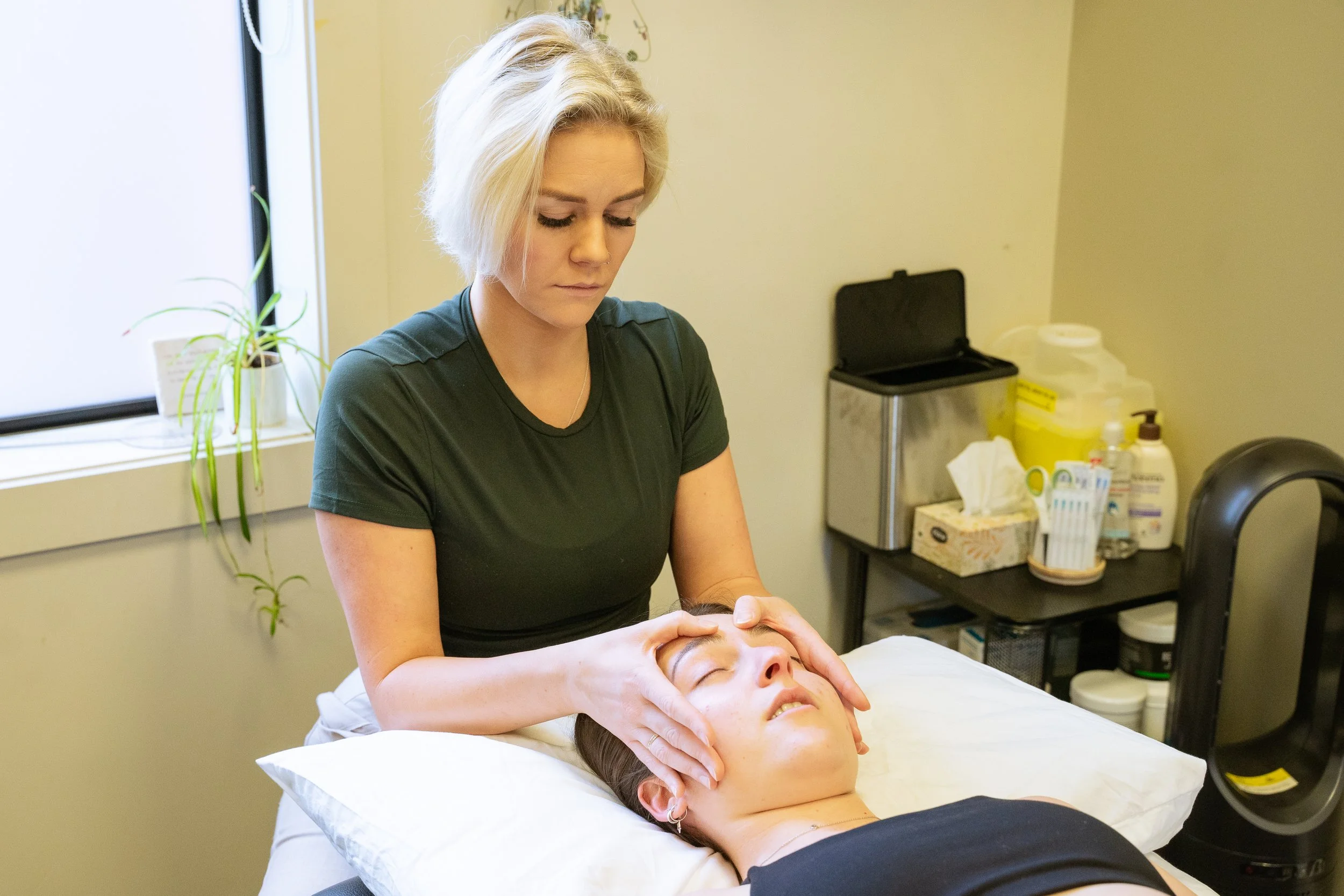 A woman receives a facial massage from a massage therapist in a treatment room with medical supplies and a window.