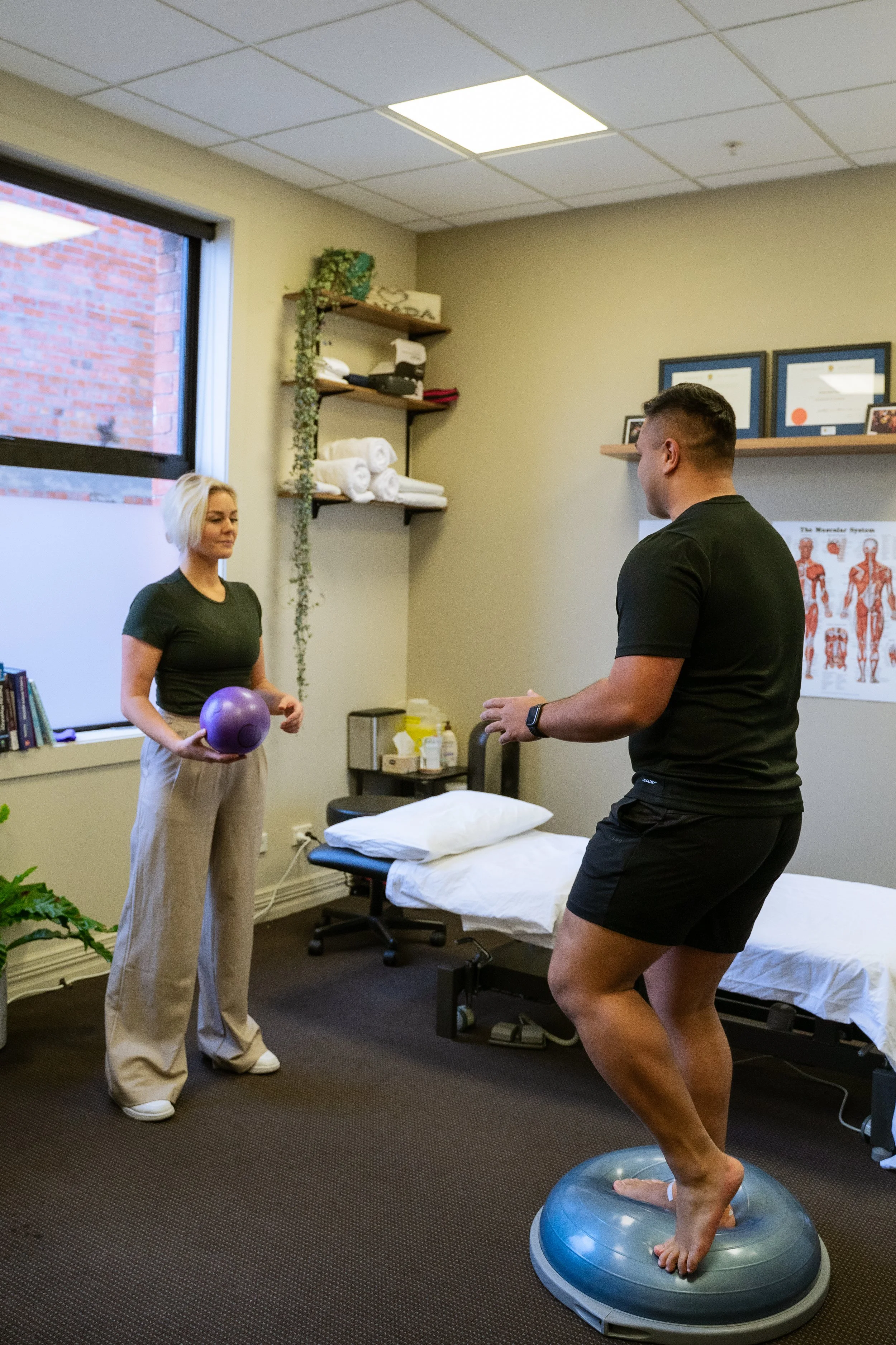 A physical therapy session in a clinic room with two individuals; one standing on a balance trainer and the other holding a purple exercise ball.