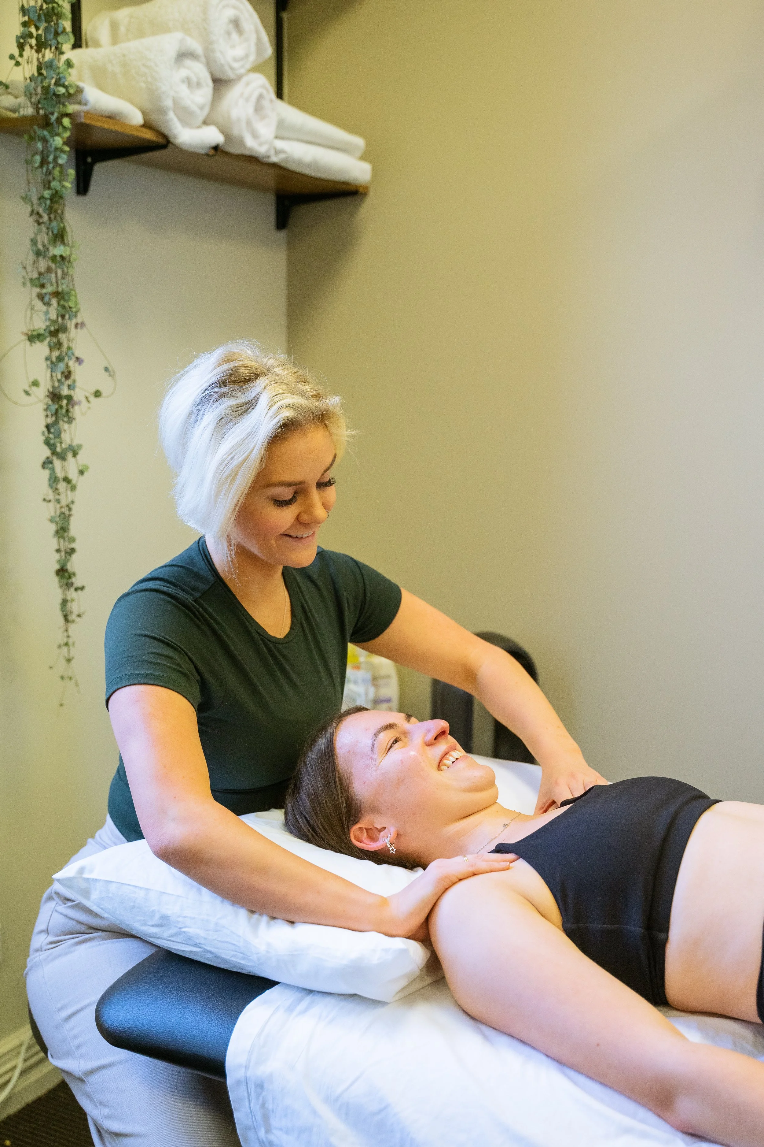 A woman lying on a massage table receiving a massage from a therapist with blonde hair, wearing a green shirt, in a softly lit room with neatly folded towels on a shelf.