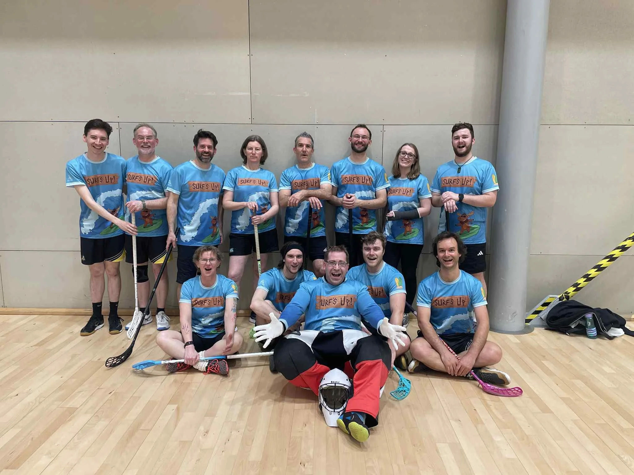 Floorball team posing in a gym wearing matching blue “Surfs Up!” jerseys, with sticks and a goalie in front.