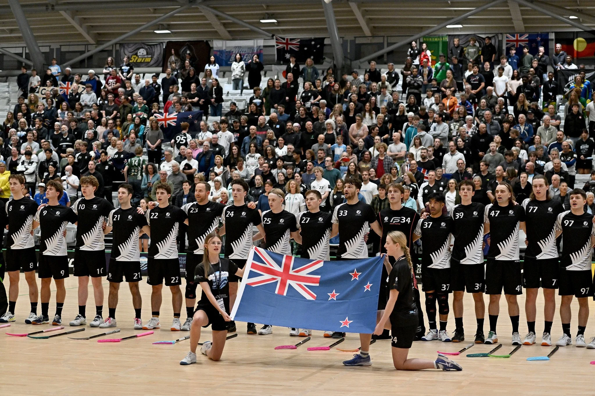 New Zealand floorball team lined up on court with a large New Zealand flag, surrounded by cheering fans.