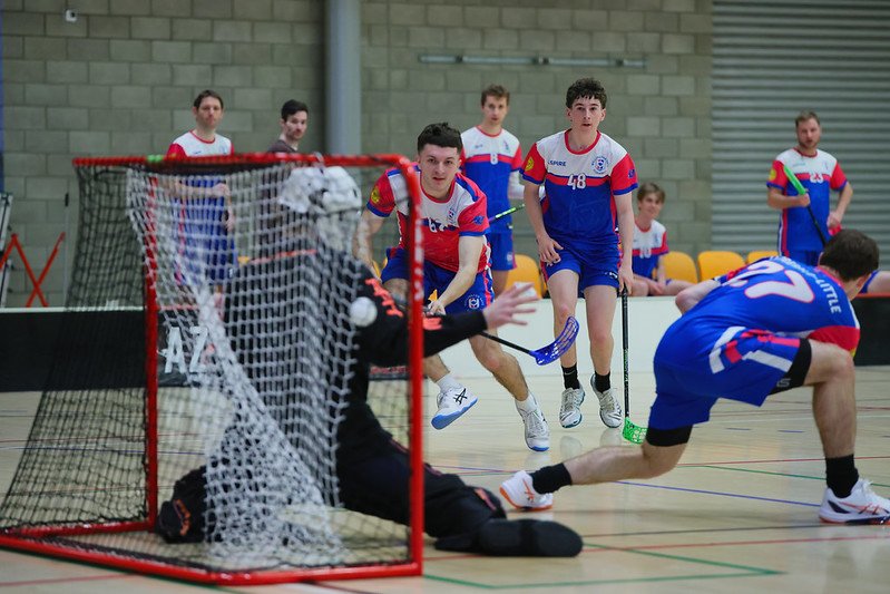 Indoor floorball players in blue and red uniforms compete near the goal as a goalkeeper blocks the ball.