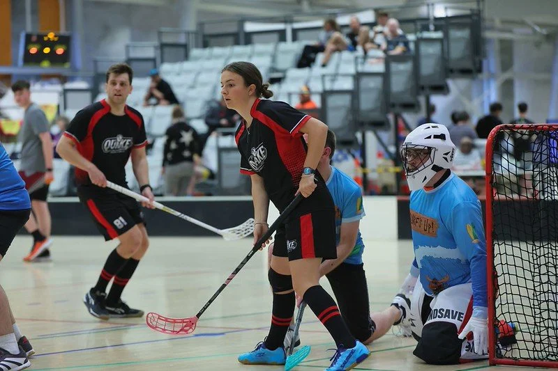 Floorball players compete near the goal during an indoor game, with a kneeling goalie in blue protective gear.
