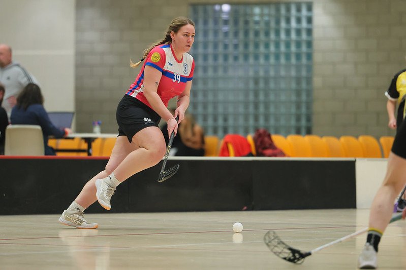 Female floorball player in a red-and-blue jersey running toward the ball with a stick during an indoor game