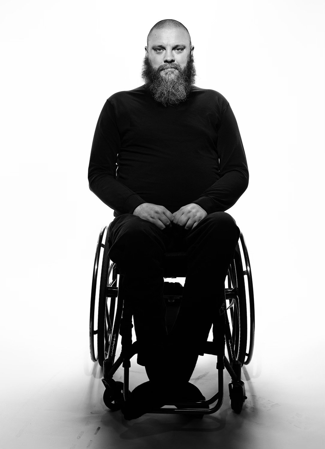 A black and white photo of a man with a buzz cut and thick beard, sitting in a wheelchair against a plain white background.