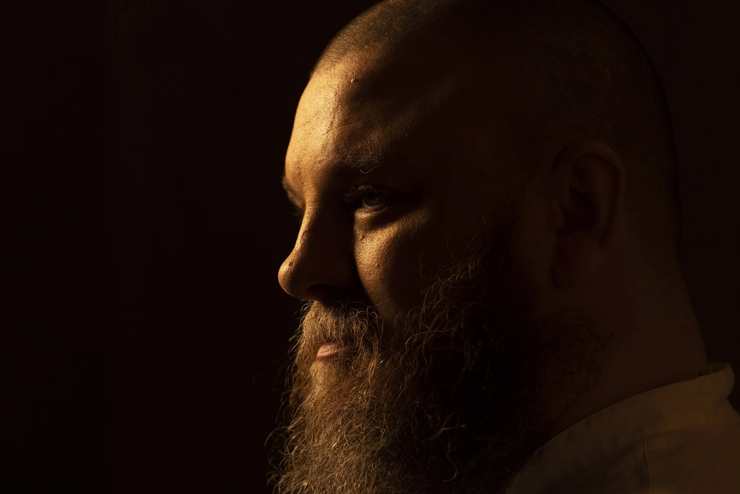 Close-up profile of a bald man with a full beard, illuminated from the left side with warm light against a dark background.