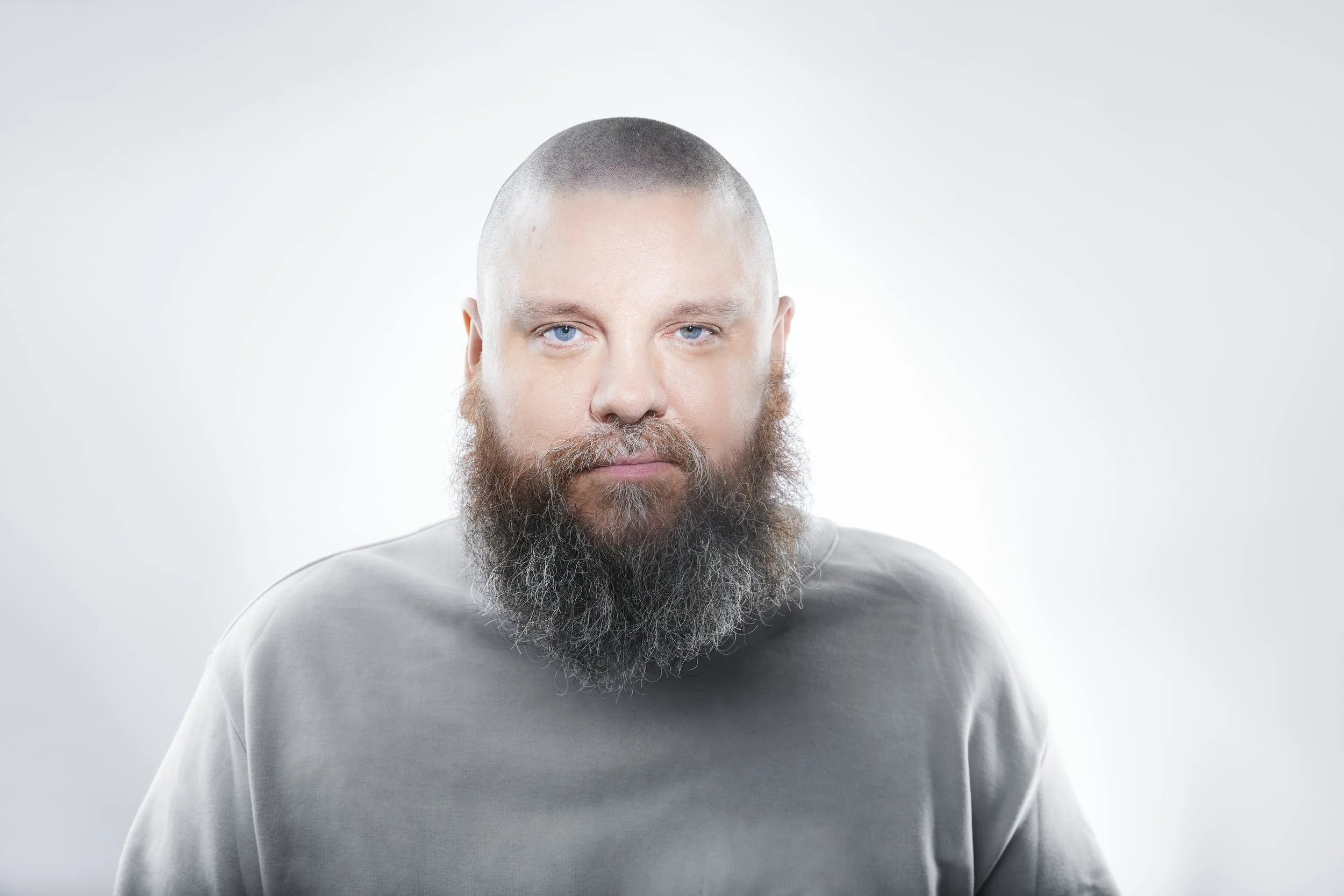A man with a shaved head, blue eyes, and a full, bushy beard wearing a light gray shirt against a plain white background.