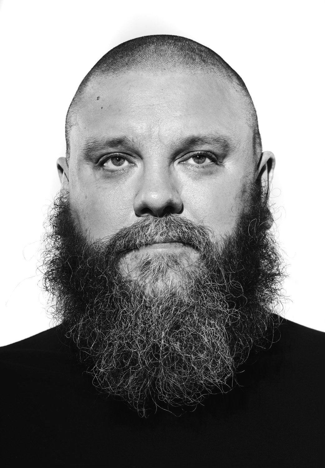 Black and white close-up portrait of a man with a short buzz haircut and a full beard, wearing a dark shirt, against a plain background.