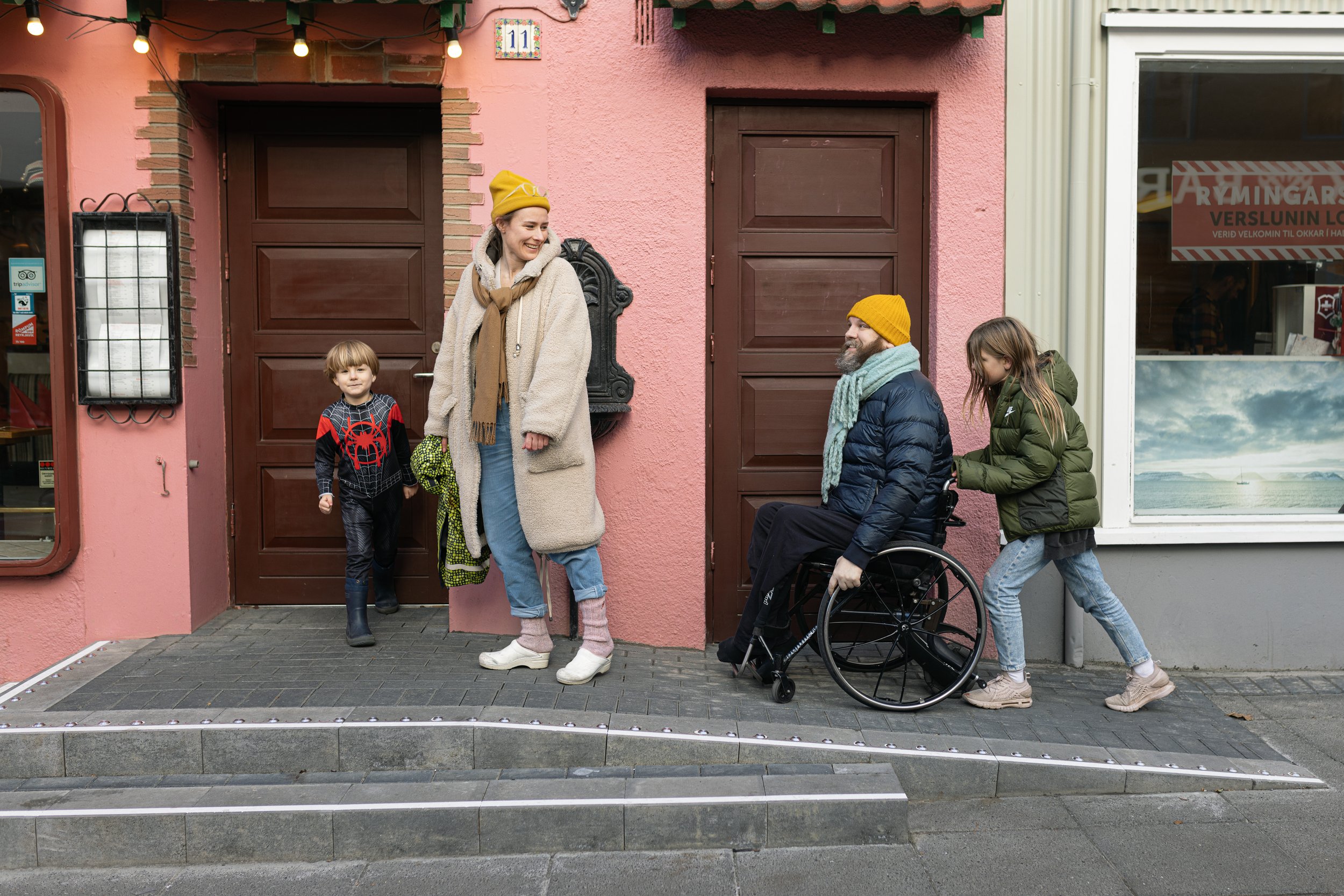 A diverse group of five people, including two children, a woman, and a man in a wheelchair, standing outside a pink building with brown doors, smiling and engaging in conversation on a sidewalk with a tactile paving strip.