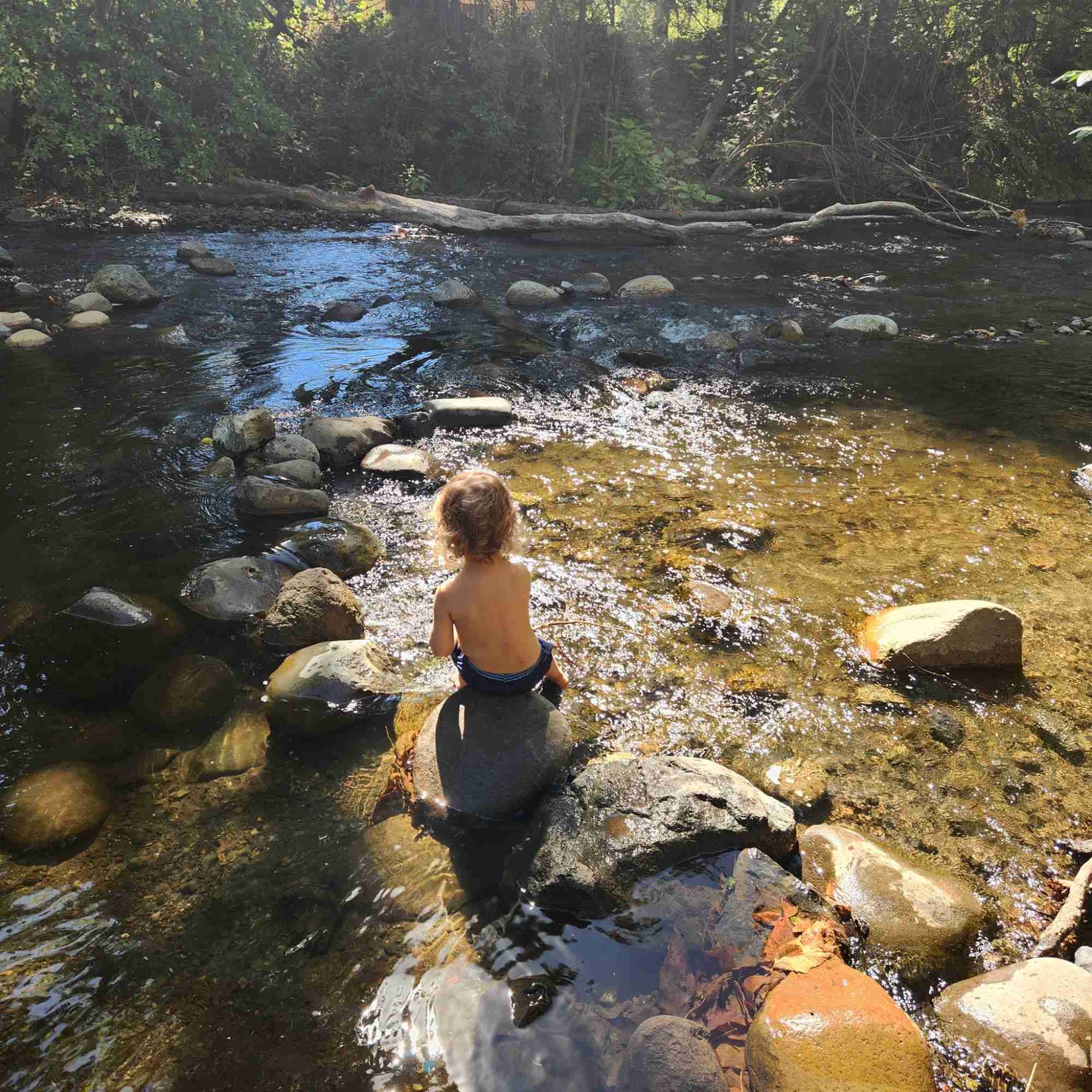 A young child at Mighty Oak Nature School with curly hair, shirtless, sitting on a large rock in a shallow river, surrounded by rocks and trees, with sunlight reflecting off the water.