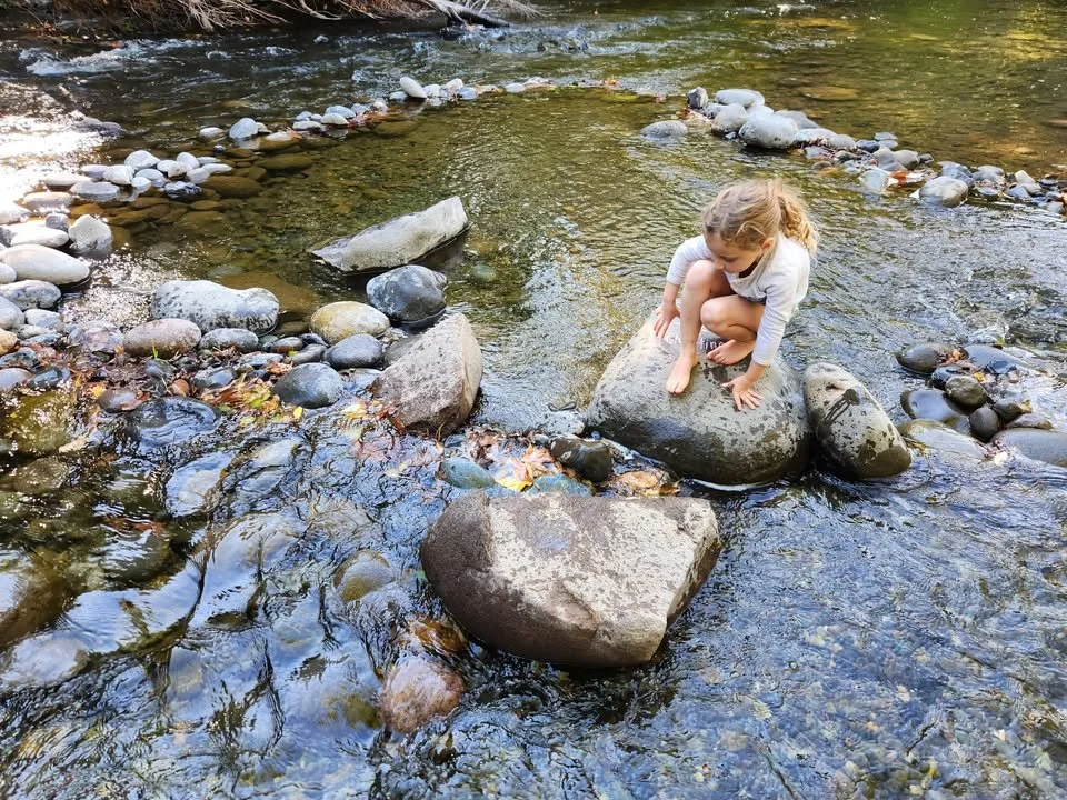 A young girl with blonde hair in a ponytail crouches on a large rock in a shallow river, surrounded by smaller rocks and pebbles, with trees reflected in the water at Mighty Oak Nature School.