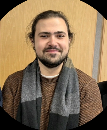 A man with long hair and a beard, wearing a brown sweater and a gray scarf, sitting in front of a wooden panel background.