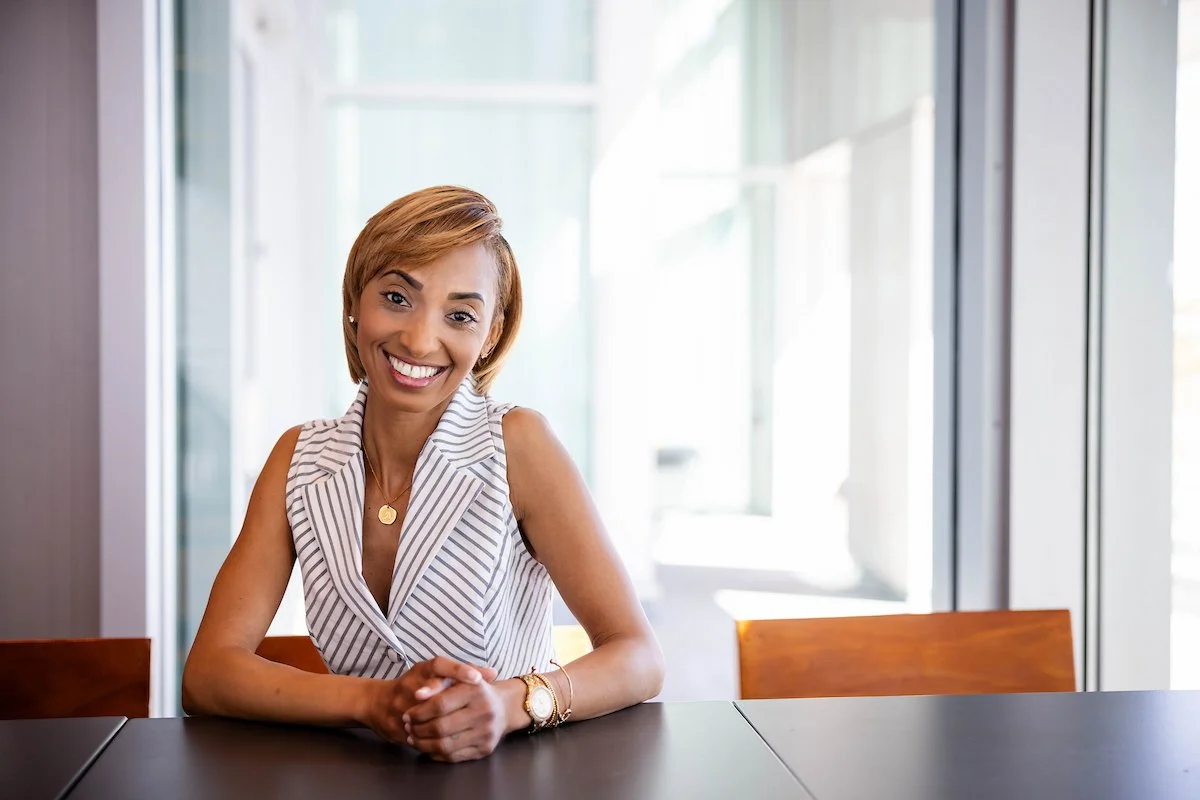 A smiling woman with short blond hair sitting at a dark table in a bright, modern office or conference room with large windows.