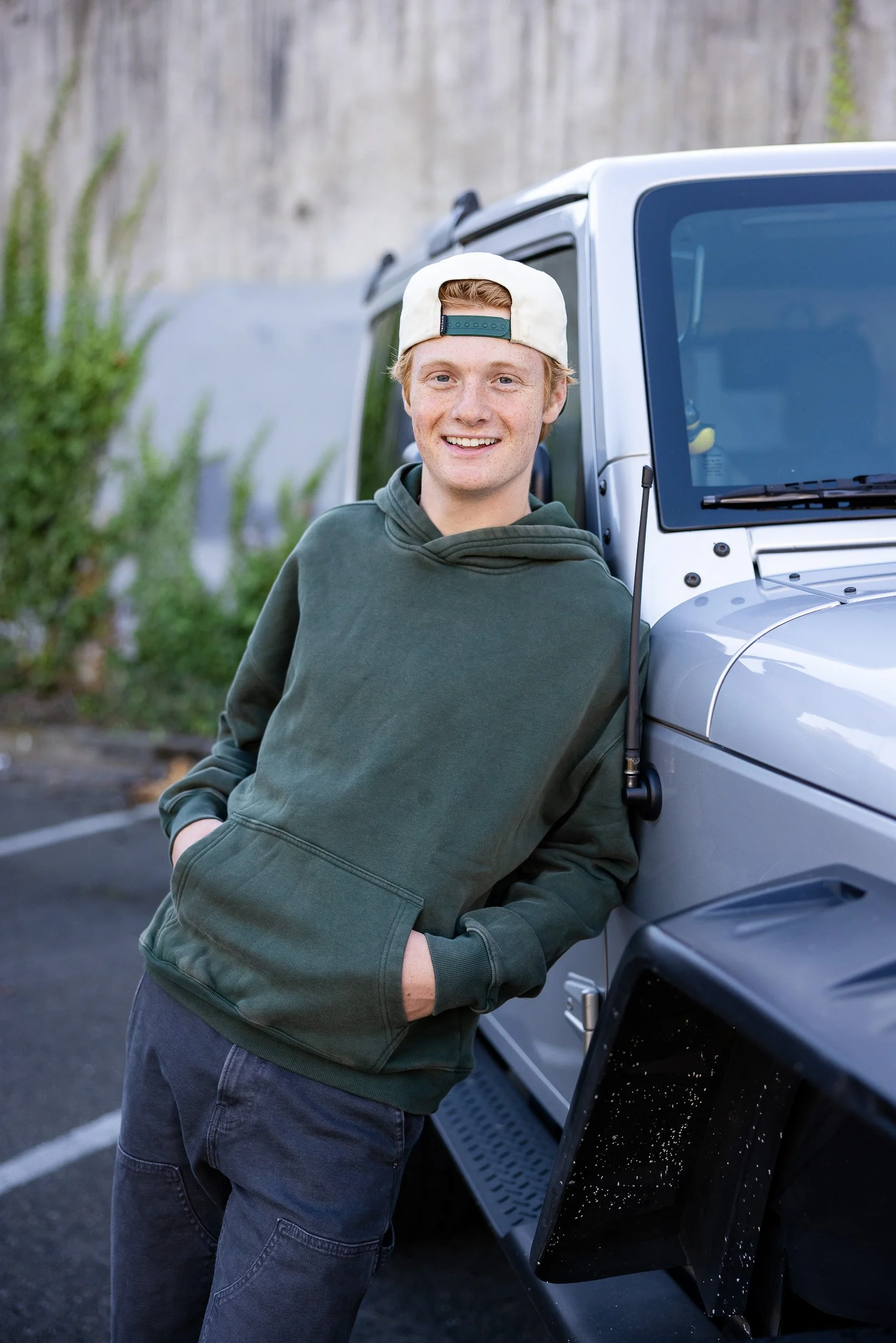 Senior portrait for high school grad. A young man wearing a green hoodie and a white backwards cap, smiling and leaning against a silver off-road vehicle, in an outdoor parking lot.
