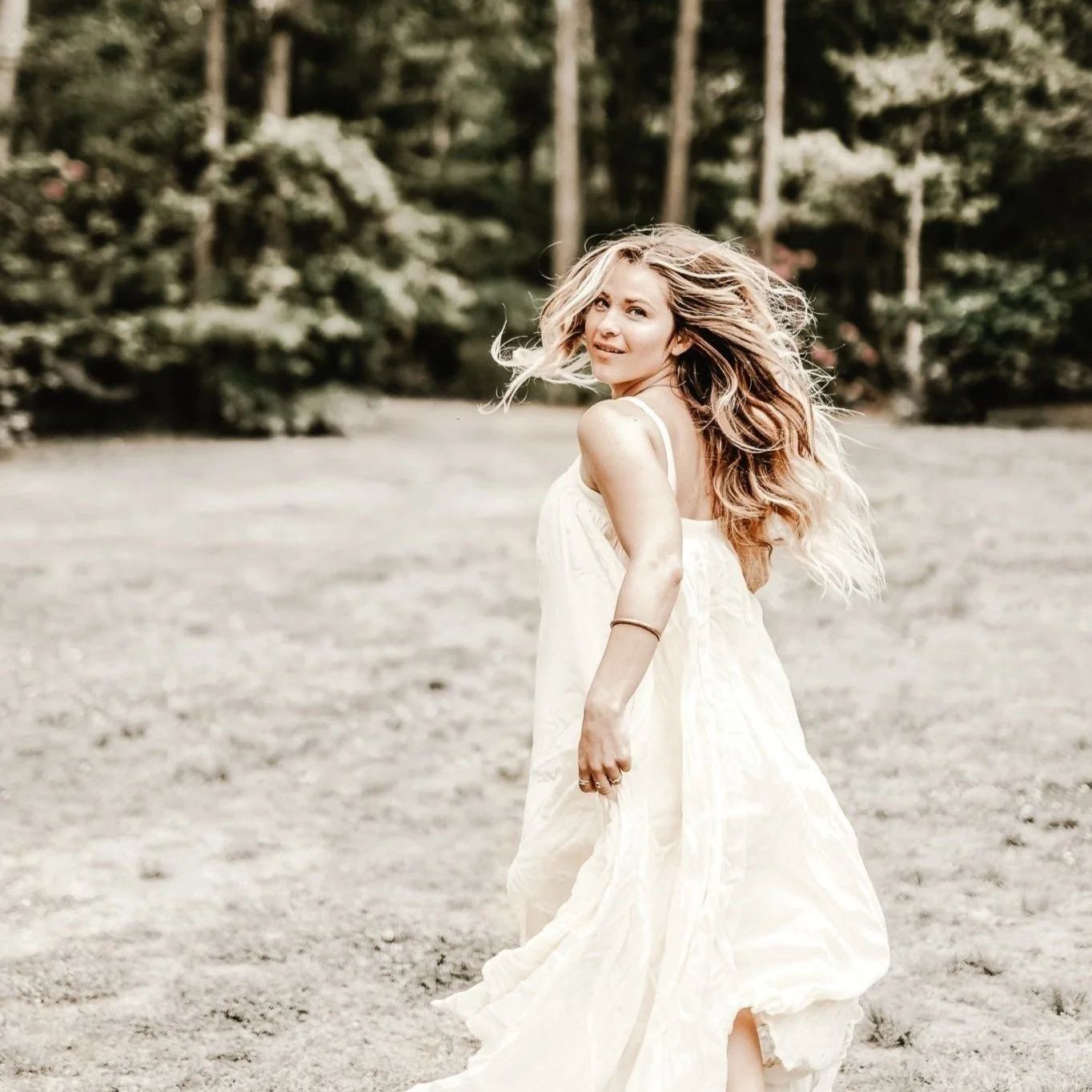 A woman with long, wavy blonde hair wearing a flowing white dress standing outdoors on a sandy surface with trees in the background, looking back over her shoulder and smiling.