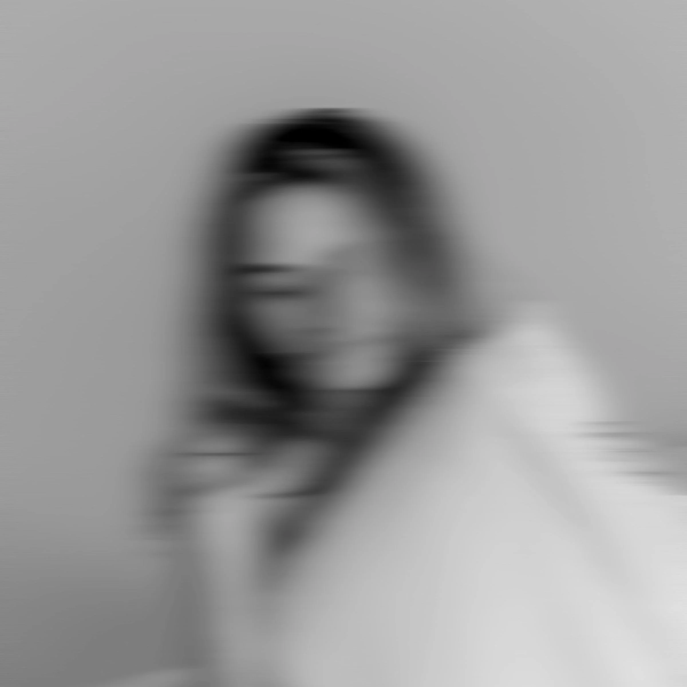 A blurred black and white photo of a woman smiling, with long dark hair, against a plain background.