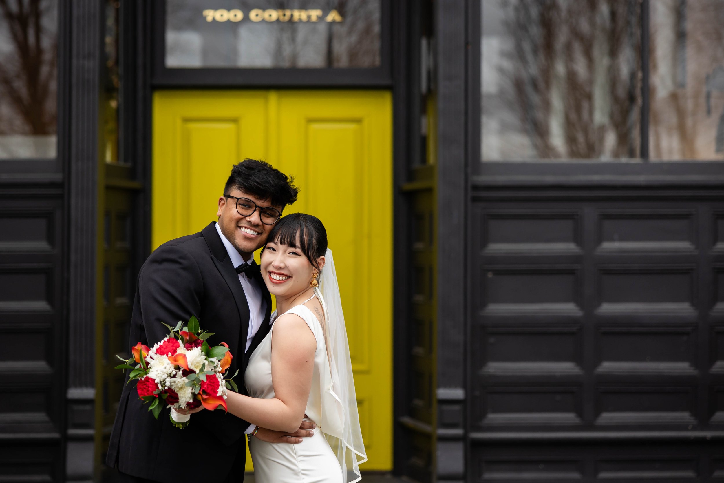 A newlywed couple smiling and hugging in front of a yellow door at Elope 253 in downtown Tacoma. The groom dressed in a black tuxedo and the bride in a white wedding gown holding a colorful bouquet.