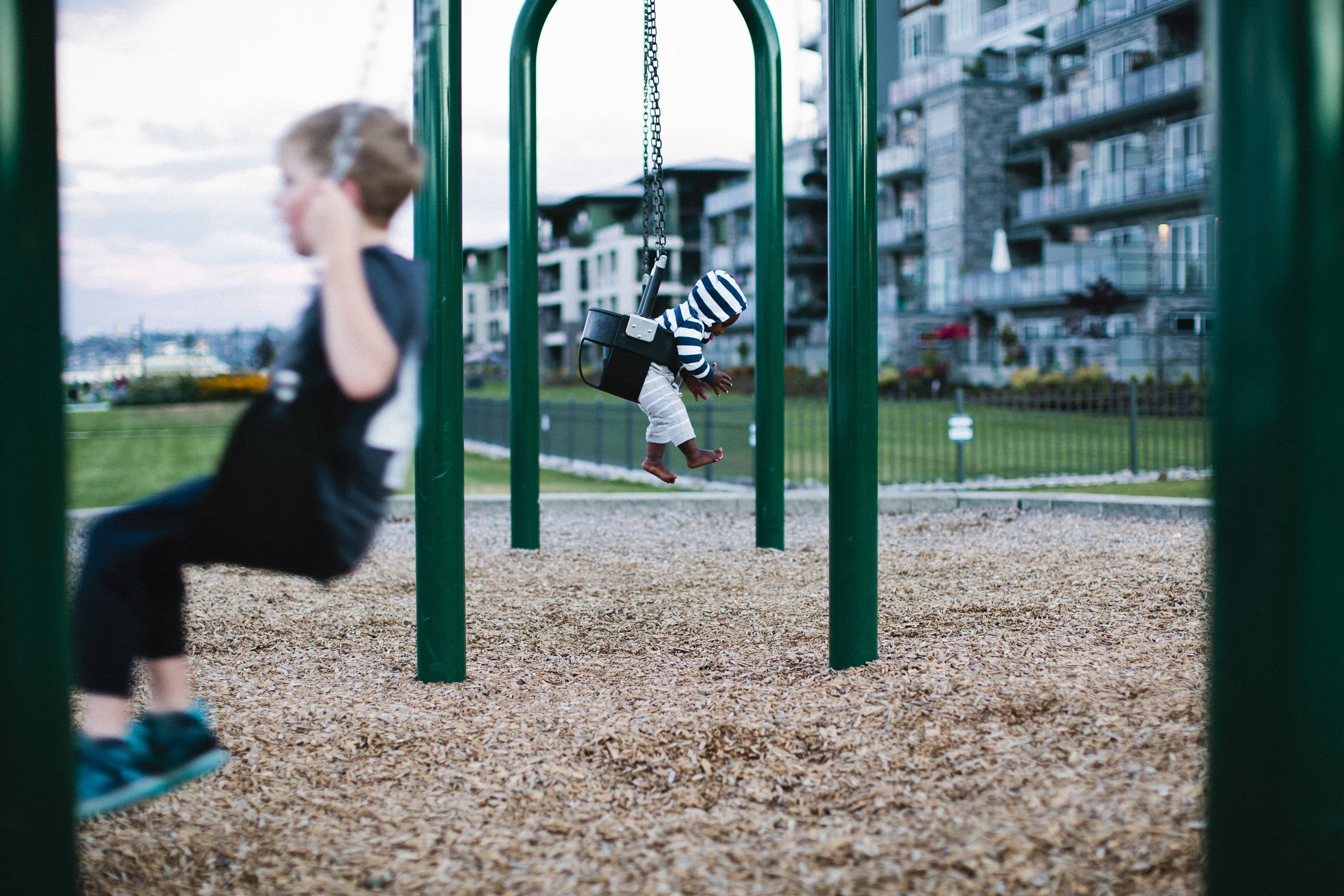 Two young children playing on swings at a park, with a background of modern apartment buildings and a cloudy sky.