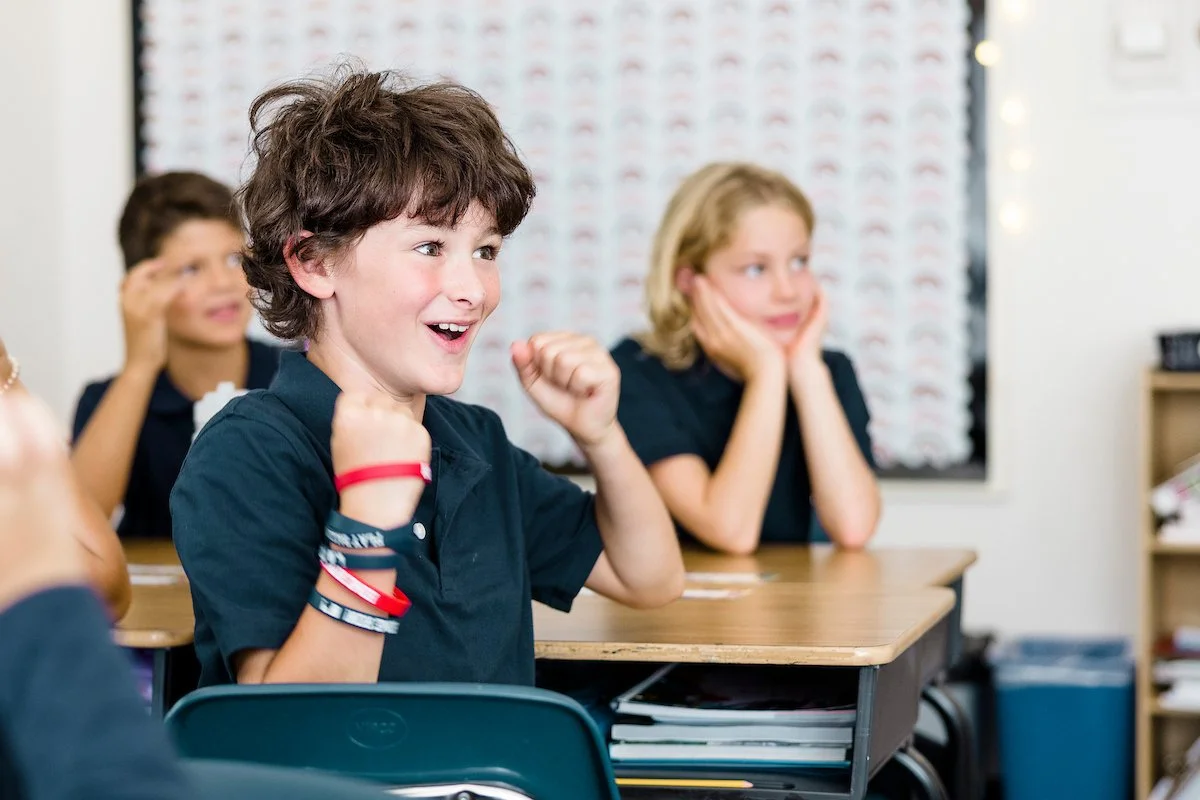 Excited school children sitting at desks in classroom, smiling, with one boy raising his fists in celebration.