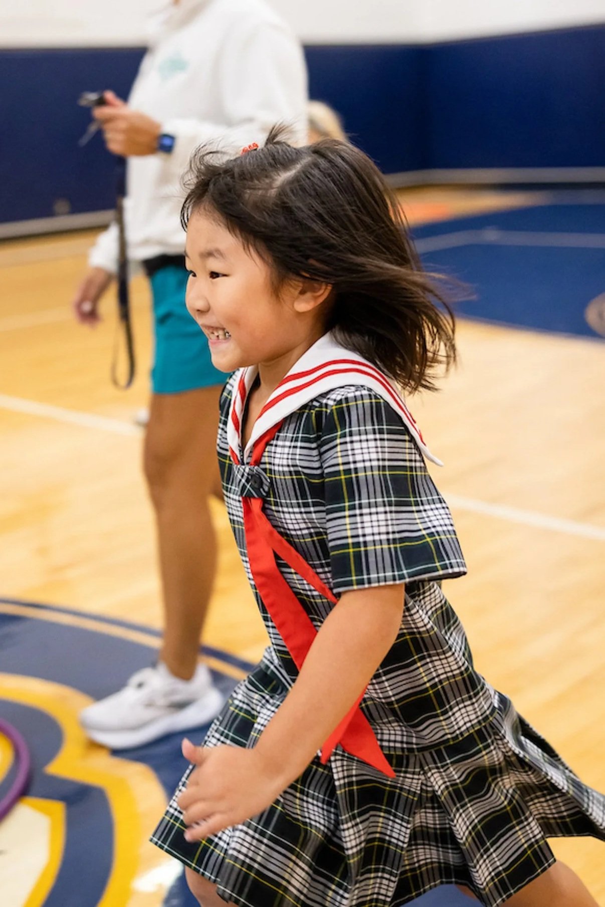 A young girl in a school uniform, smiling and running indoors with windblown hair. In the background, a person holds a camera and wears casual clothes.