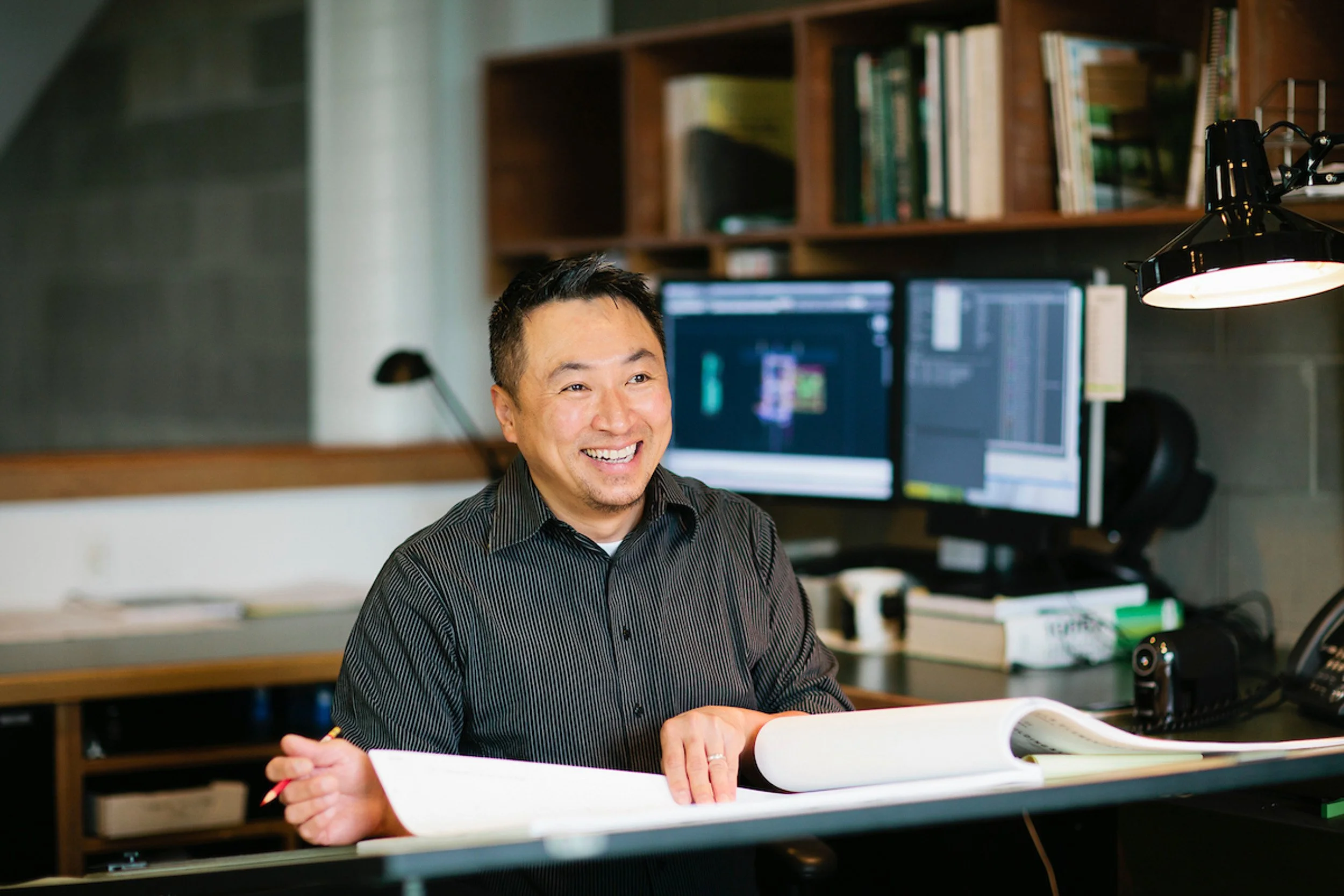 A smiling man sitting at a desk with an open notebook, in front of two computer monitors, in a modern office or studio setting.
