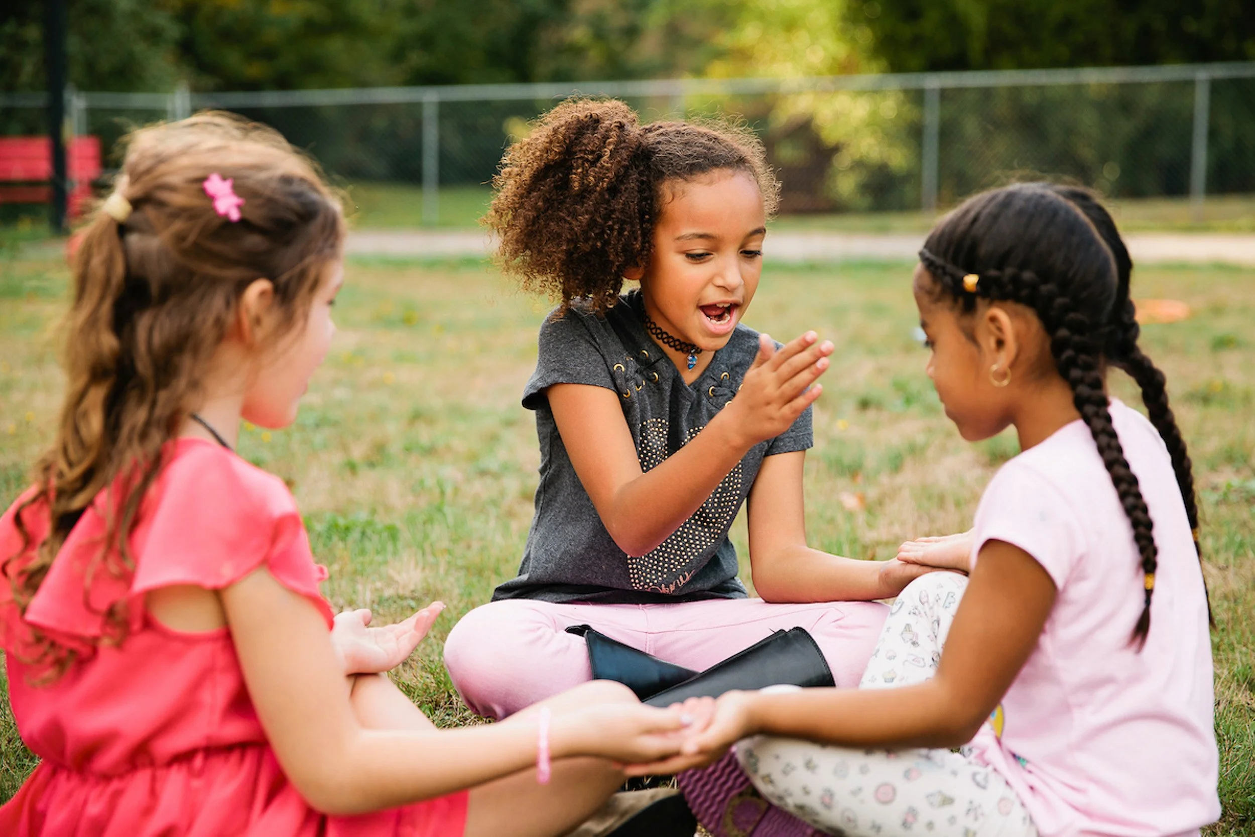 Three girls sitting on the grass outdoors, engaging in a game or activity, with a fence and trees in the background.