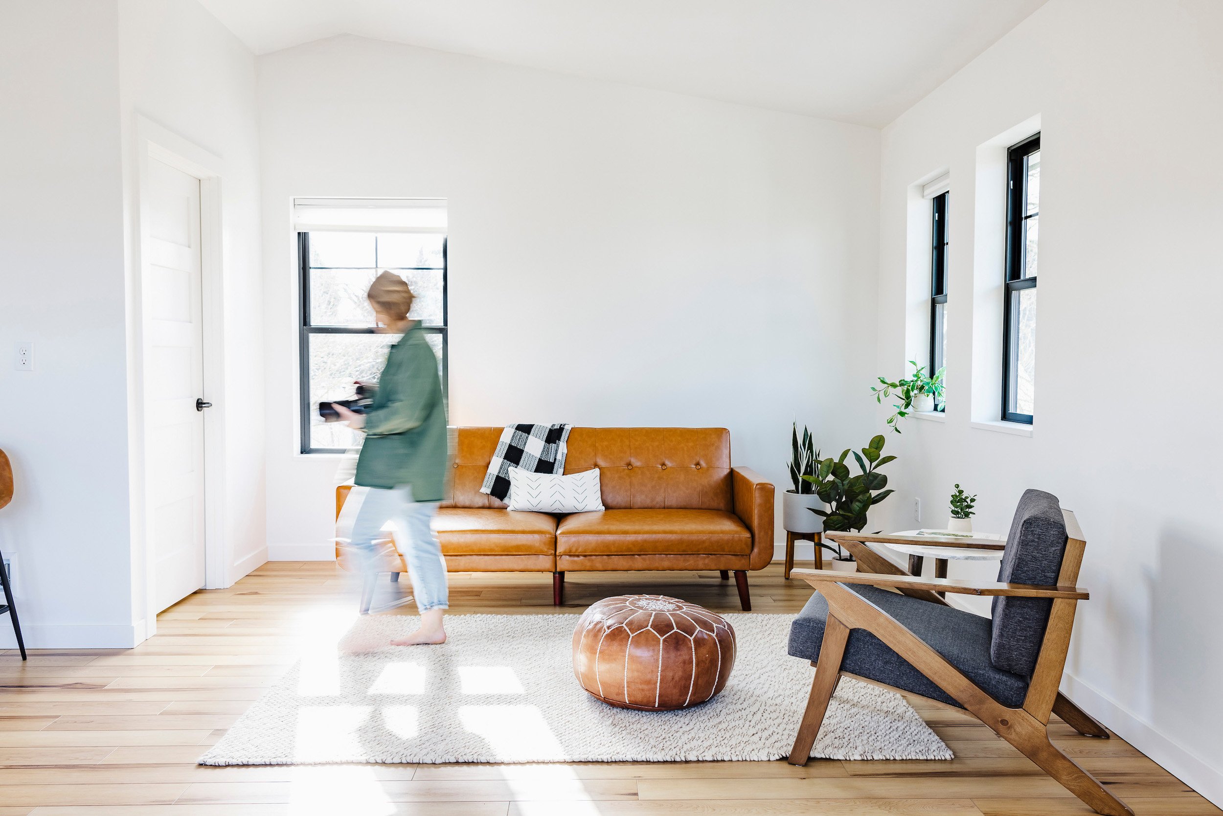 Living room with white walls, wooden floor, tan leather sofa, wooden armchair, green plants, and a person walking past holding an object.