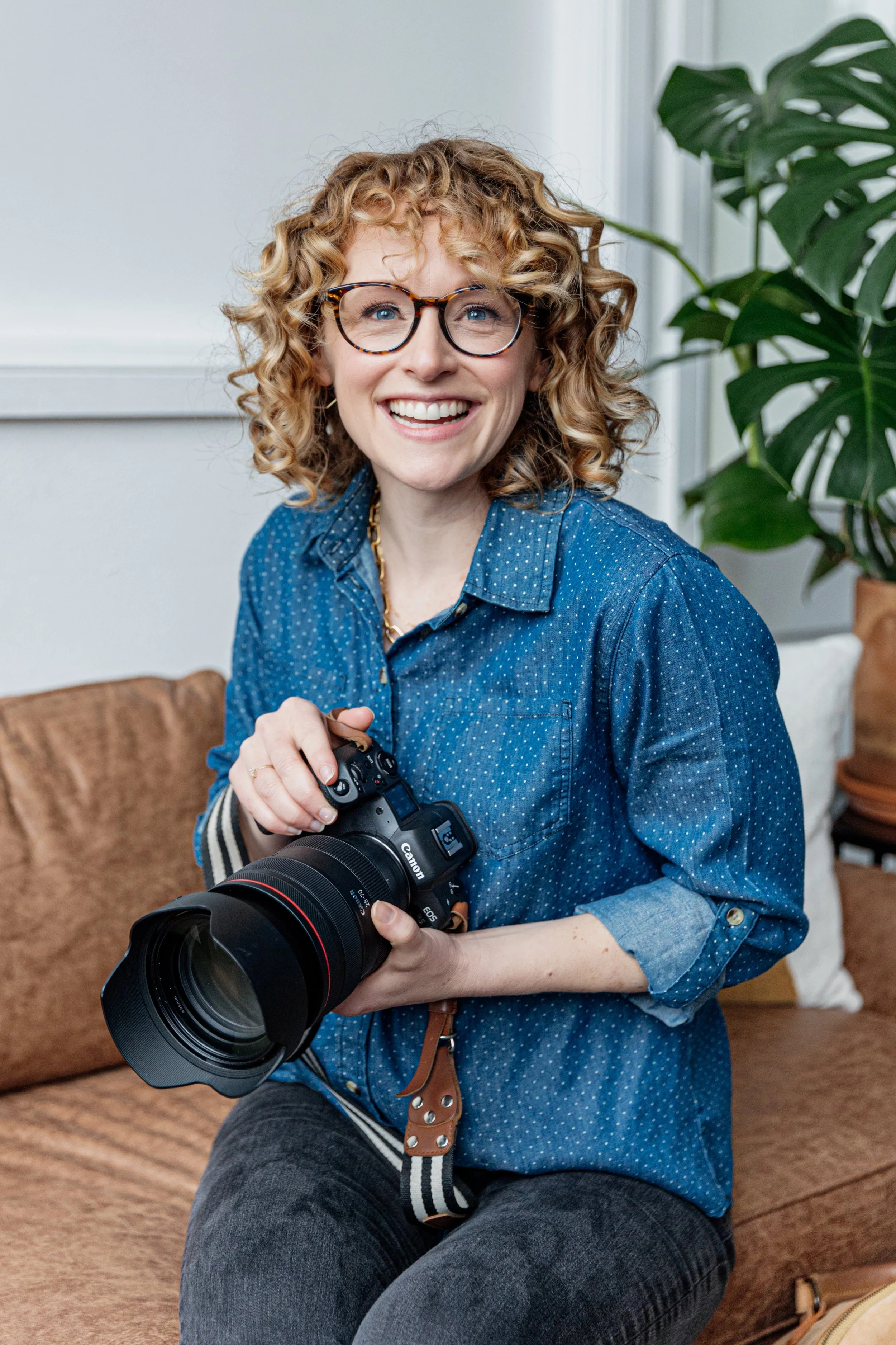 A woman with curly blonde hair, glasses, and a blue polka-dot shirt, sitting on a brown couch, holding a professional camera with a large lens, smiling and looking happy.