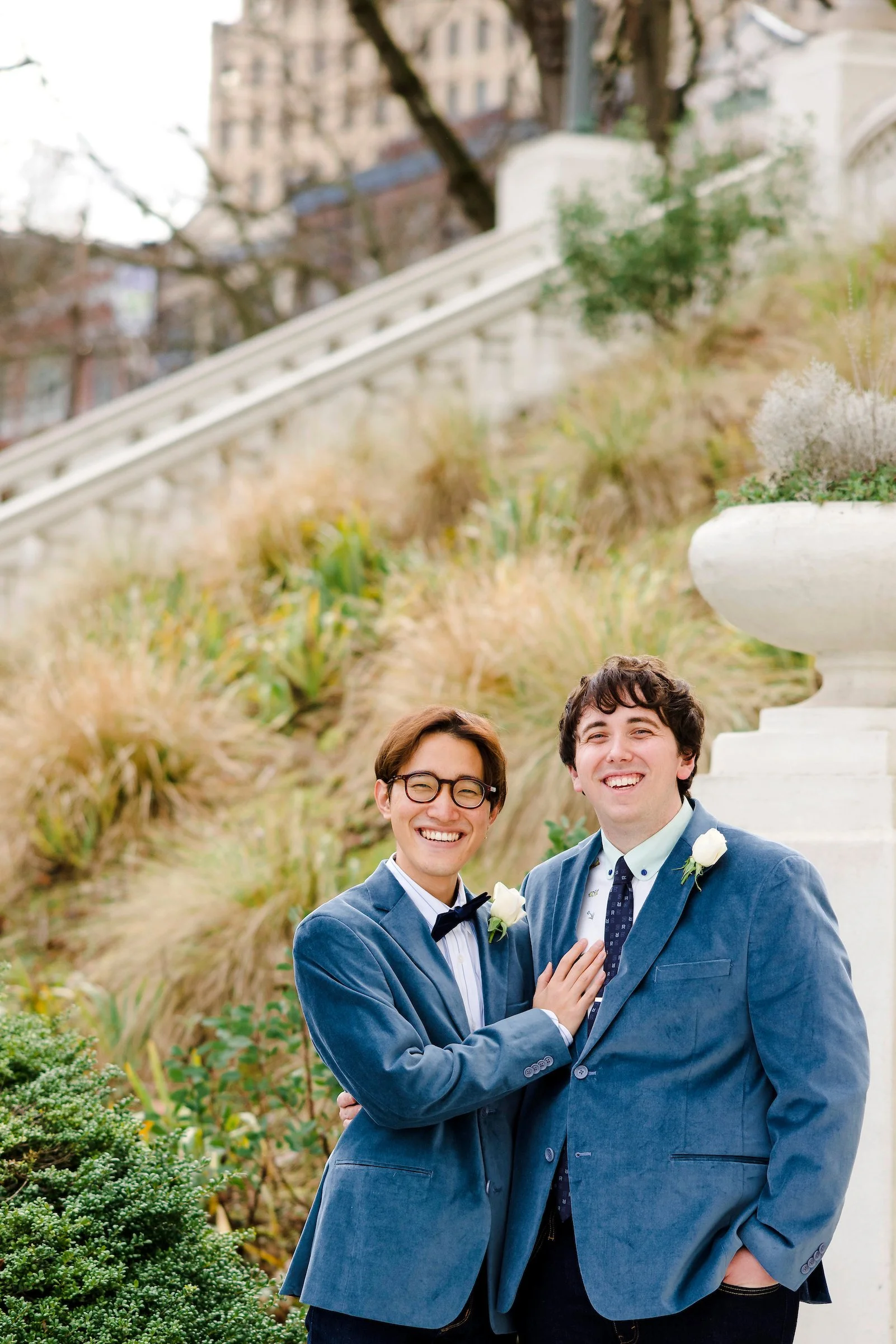 Two men dressed in blue suits with white shirts and dark ties, standing outdoors in front of a white decorative urn, smiling at the camera, with a garden and city buildings in the background.
