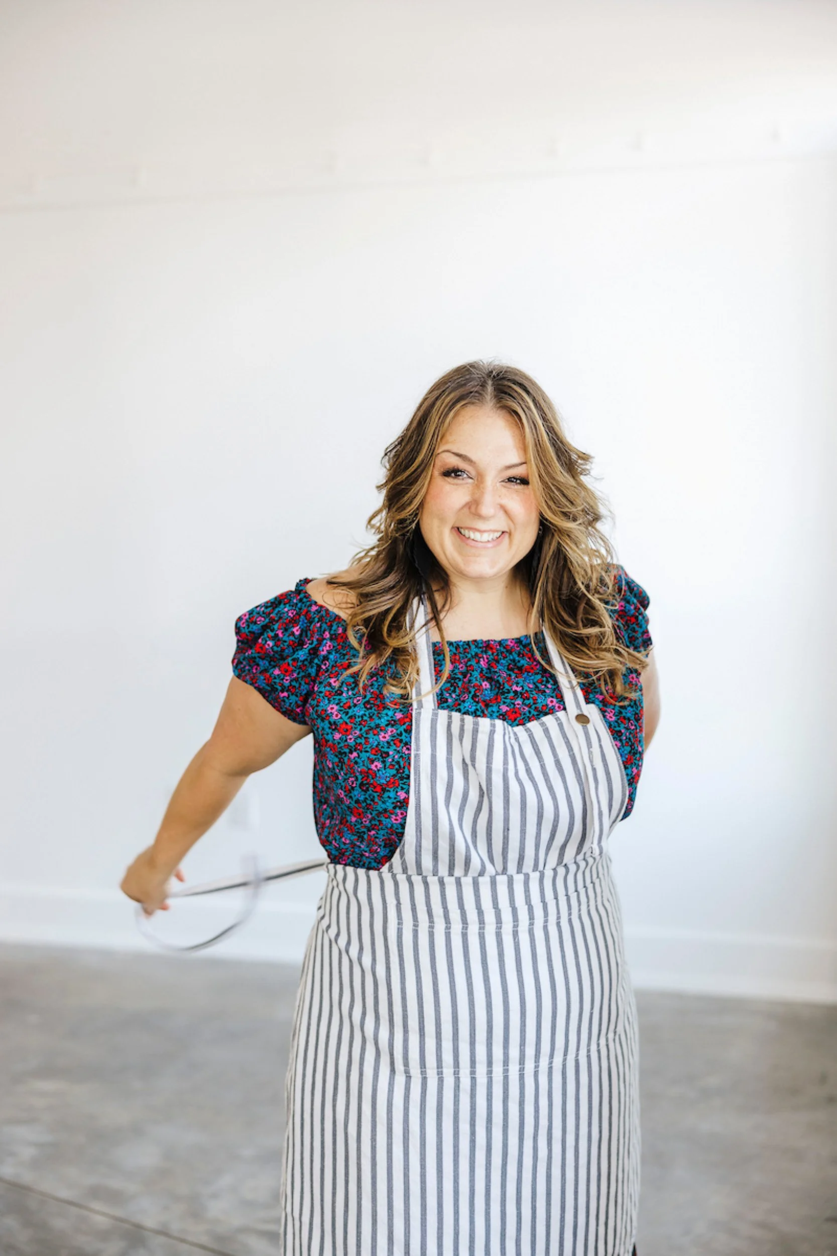 A woman with long wavy hair smiling and winking, wearing a colorful floral top and a striped apron, standing in a bright, minimalistic room.