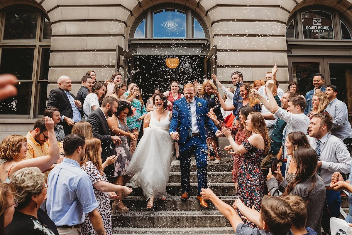 Wedding celebration with the bride and groom walking down the steps outside a courthouse, surrounded by friends and family throwing confetti and cheering.