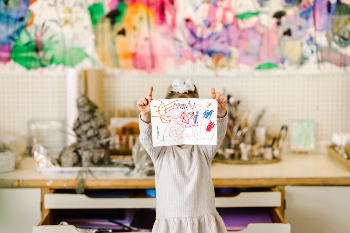 Child holding up a colorful crayon drawing in an art studio.