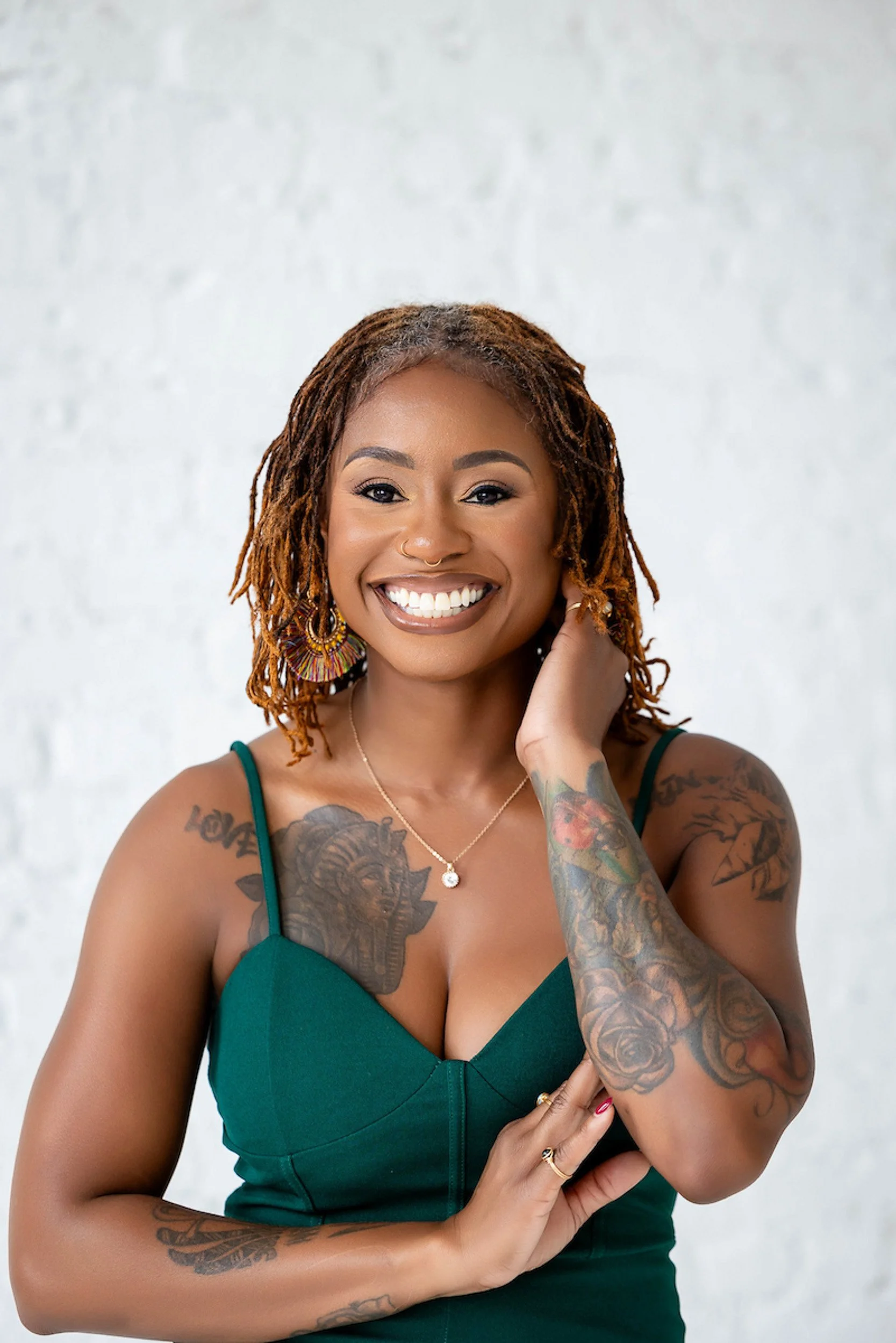 A smiling woman with dreadlocks and tattoos, wearing a green dress and colorful earrings, posing against a white background.