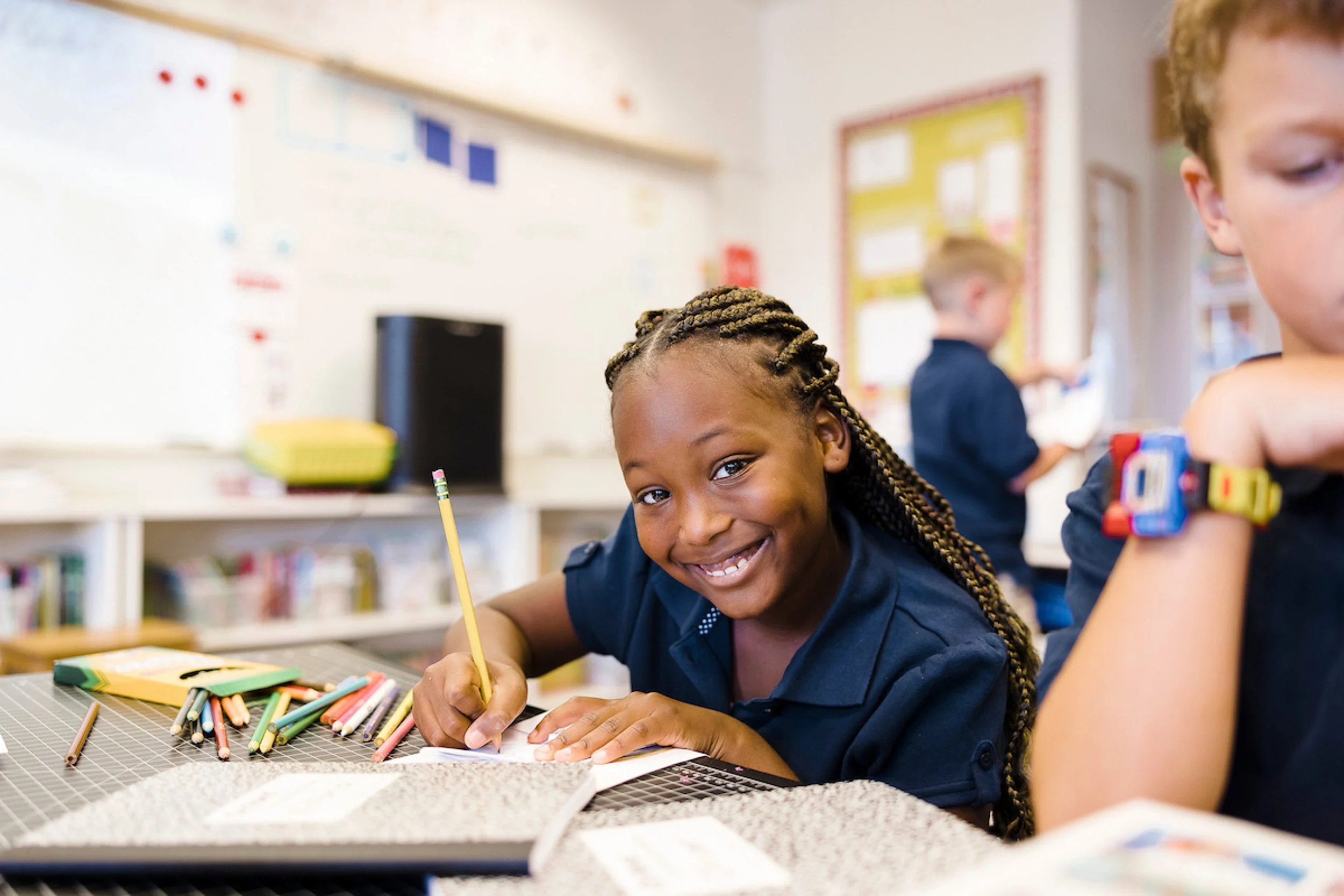 Smiling girl with braided hair writing in a notebook in a classroom with school supplies on the table.