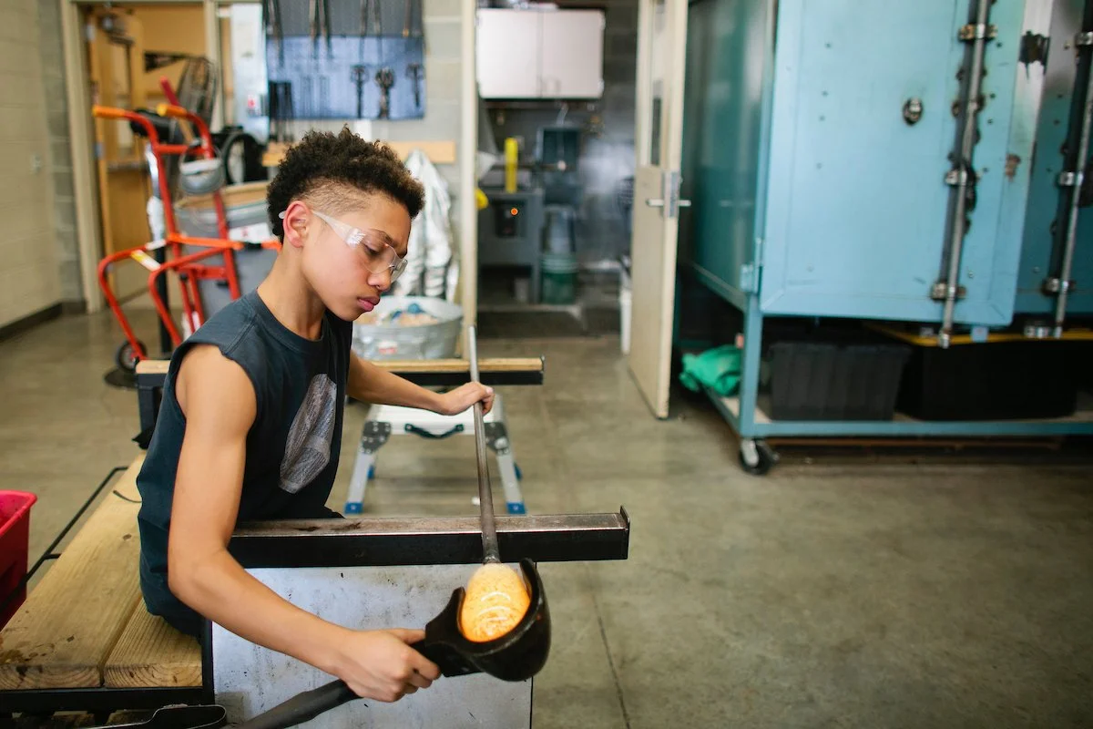 A boy working in a glass blowing workshop with a nonprofit called Hilltop Artists Hilltop Heritage Middle School in Tacoma WA. He is wearing safety glasses and a sleeveless shirt. The workshop has equipment, storage cabinets, and a gray floor.