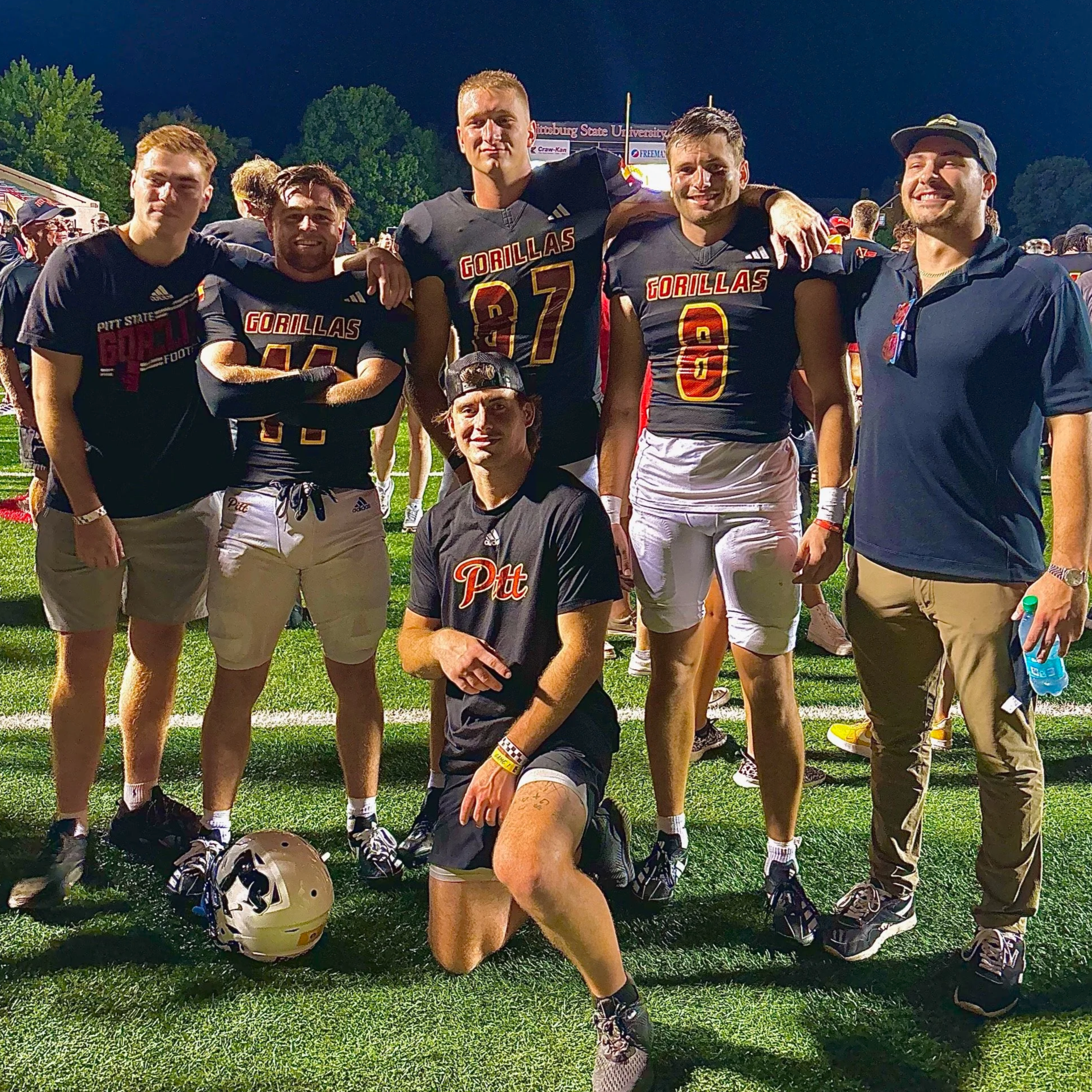 A group of football players and a coach pose for a photo on a grassy field at night. The players are wearing black jerseys with "Gorillas" written on them and have helmets on the ground nearby. They are smiling with their arms around each other. The background shows a crowd of spectators and a scoreboard.