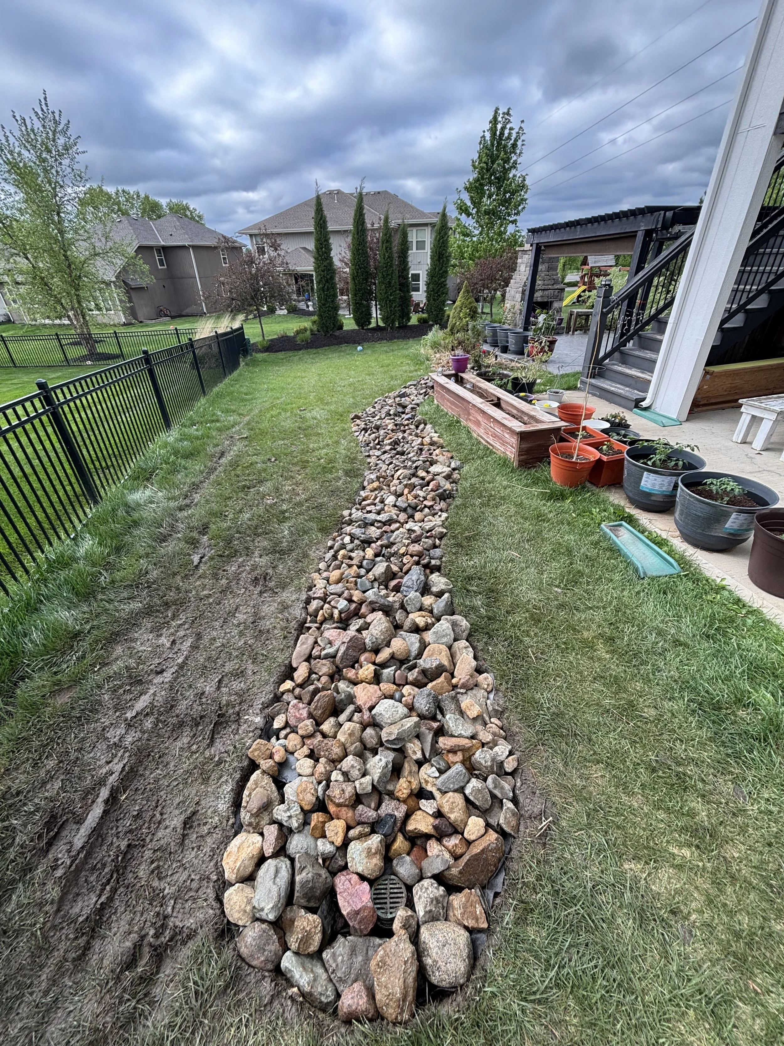A backyard with a lawn, a black metal fence, and a row of rocks forming a border. There are potted plants and garden beds beside a porch with stairs. In the background, there are trees and houses under a cloudy sky.