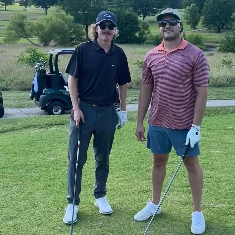 Two men standing on a golf course holding golf clubs, wearing golf gloves, sunglasses, and casual golf attire, with a golf cart and scenic greenery in the background.