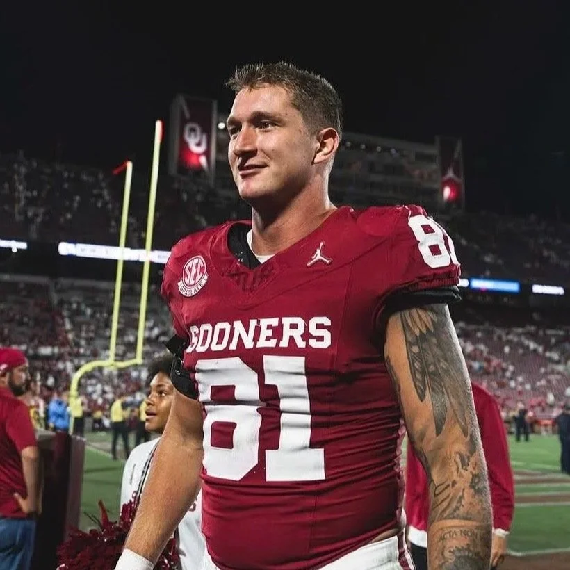 A male football player in a red Oklahoma Sooners jersey with the number 81 on it, standing on the field at night after a game, with a stadium and crowd in the background.