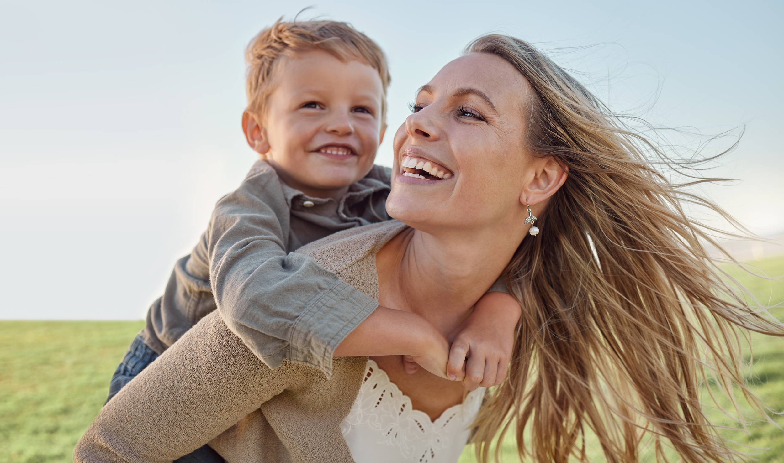 A woman with long, flowing hair carrying a young boy on her back, both smiling and enjoying a sunny outdoor setting.