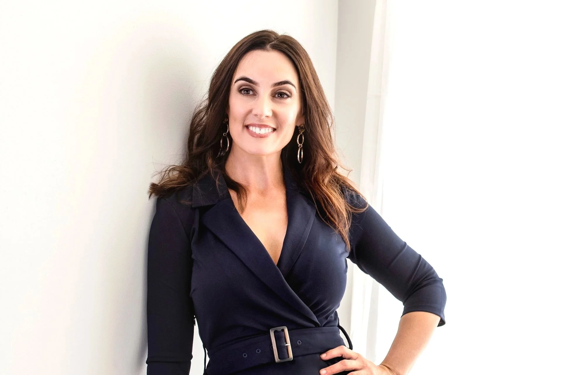 Kylie Cloney wearing a dark navy dress and earrings, standing indoors near a window with a white wall background.