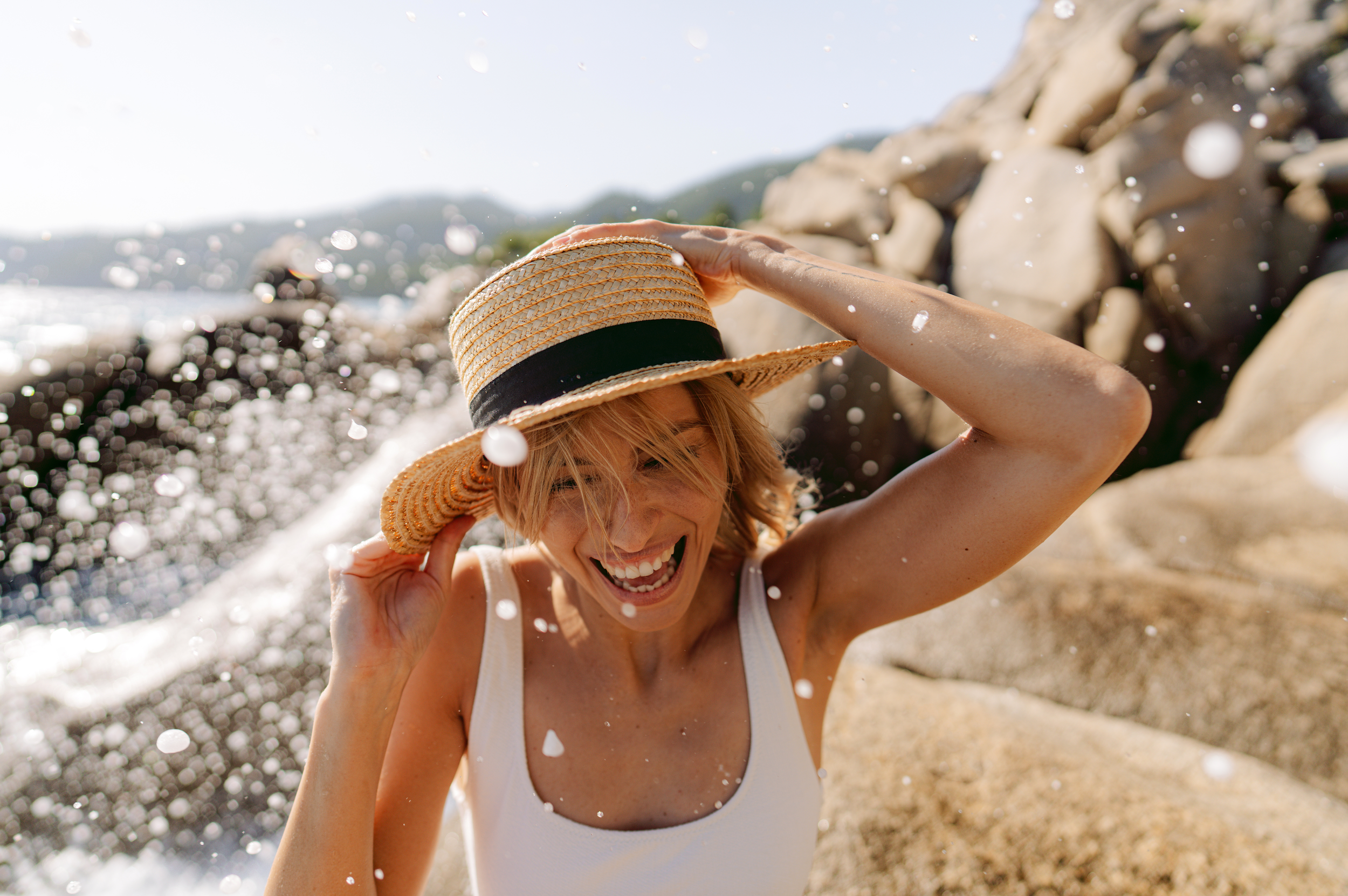 A woman smiling and wearing a straw hat with a black ribbon, enjoying a splash of water on a rocky beach on a sunny day.