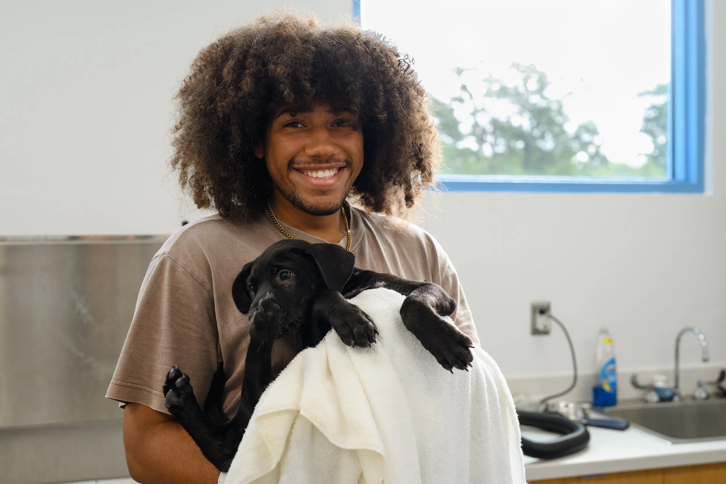 A smiling man with curly hair holding a black puppy wrapped in a white towel in a veterinary or animal care facility.