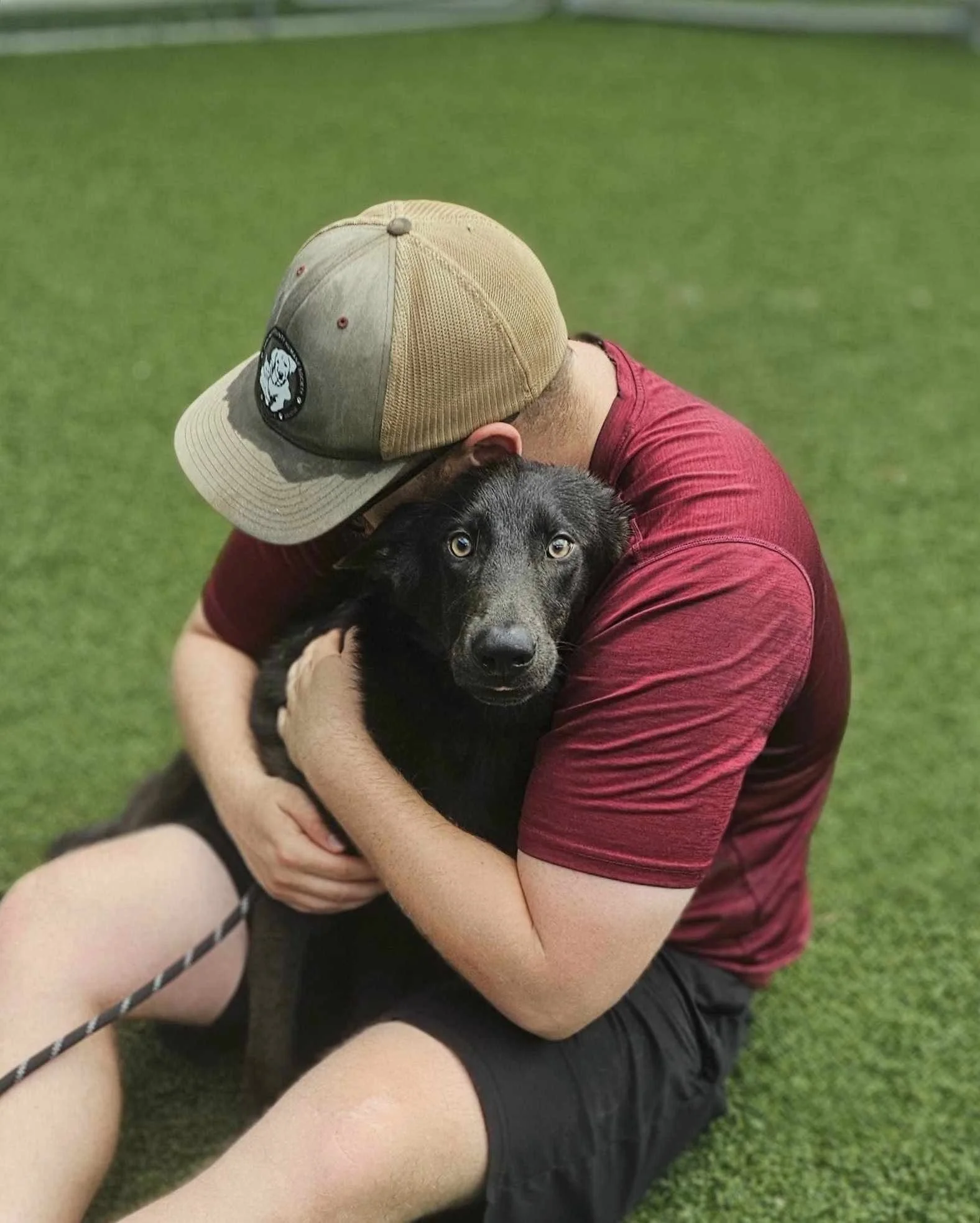A man in a red shirt and a tan cap hugging a black puppy on a grassy field.