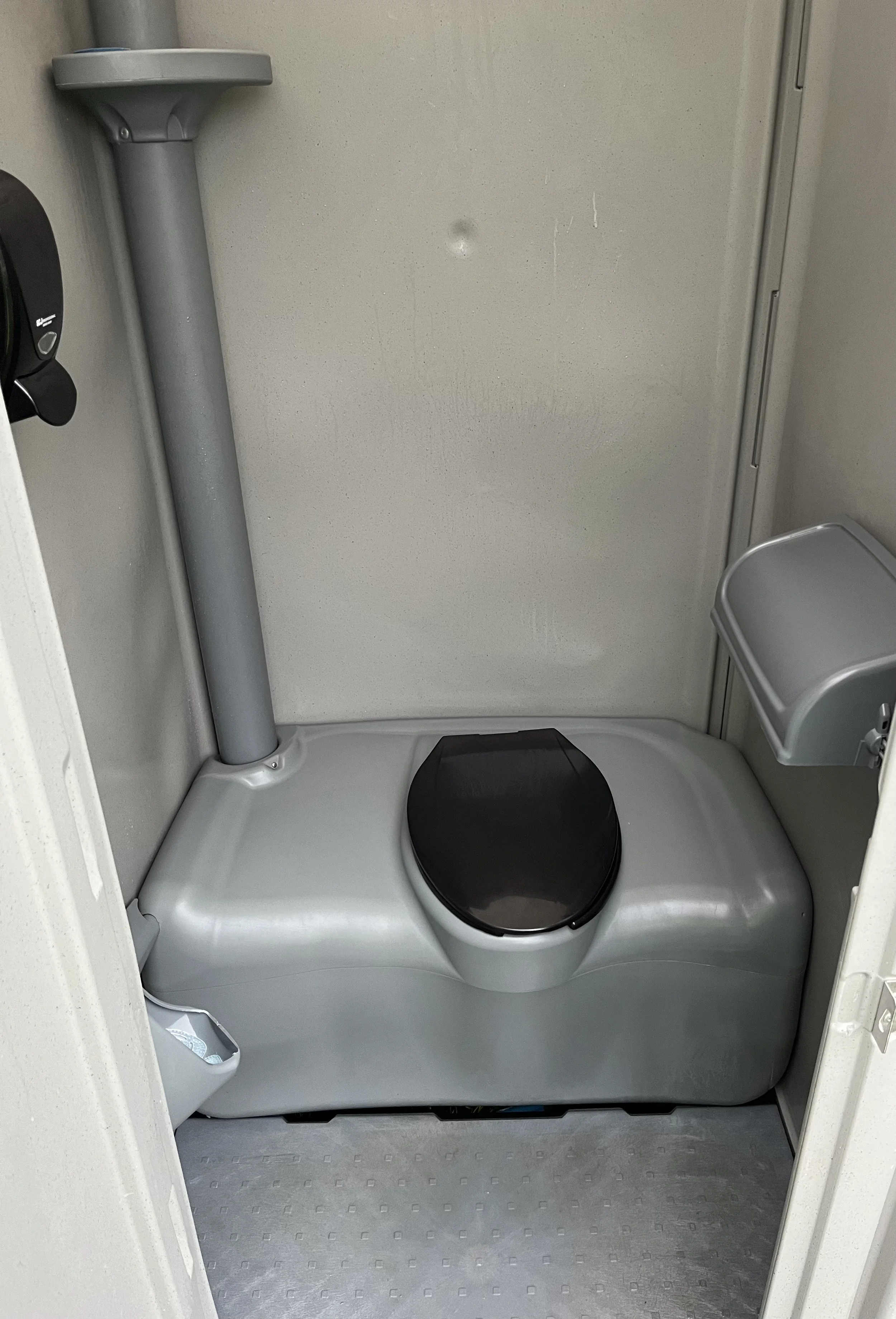 Small portable toilet with a black seat, gray walls, a small silver trash bin, and a gray hand sanitizer dispenser on the side.